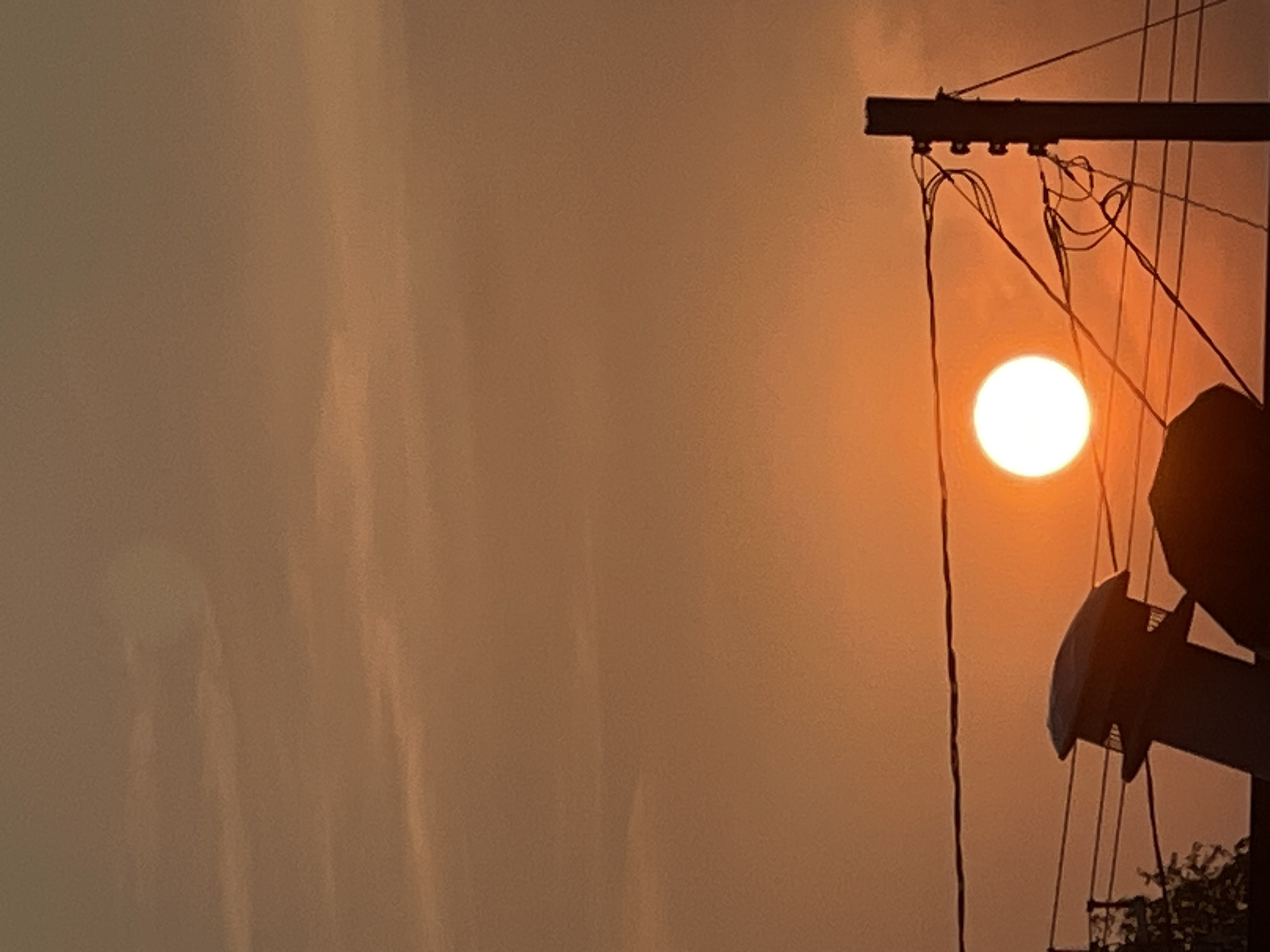 The image captures a sunset over Bluewater, Canada, characterized by a vibrant orange sky with thin, wispy clouds. The sun, a bright, luminous orb, is positioned centrally in the frame. In the foreground, silhouetted against the sunset, are several man-made structures. A power line supported by a wooden pole with insulators runs horizontally across the lower half of the image. To the left, a ventilation pipe from a building extends upwards. The scene is devoid of any people or animals, emphasizing the natural phenomenon of the setting sun and the urban infrastructure.
