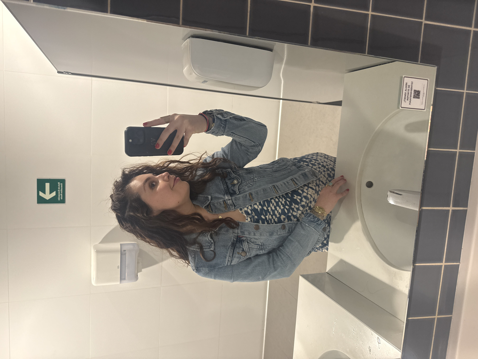 A woman with curly brown hair is taking a selfie in a bathroom. She is wearing a denim jacket over a blue and white patterned dress. She is holding a black smartphone in her left hand, with red nail polish on her fingers. The bathroom has white tiles and a sink with a silver faucet. There is a hand dryer and a green sign with a white arrow pointing down on the wall. There is a sticker with text in Spanish and a QR code on the upper right corner of the mirror. The location of the photo is likely in Mexico City, Mexico.