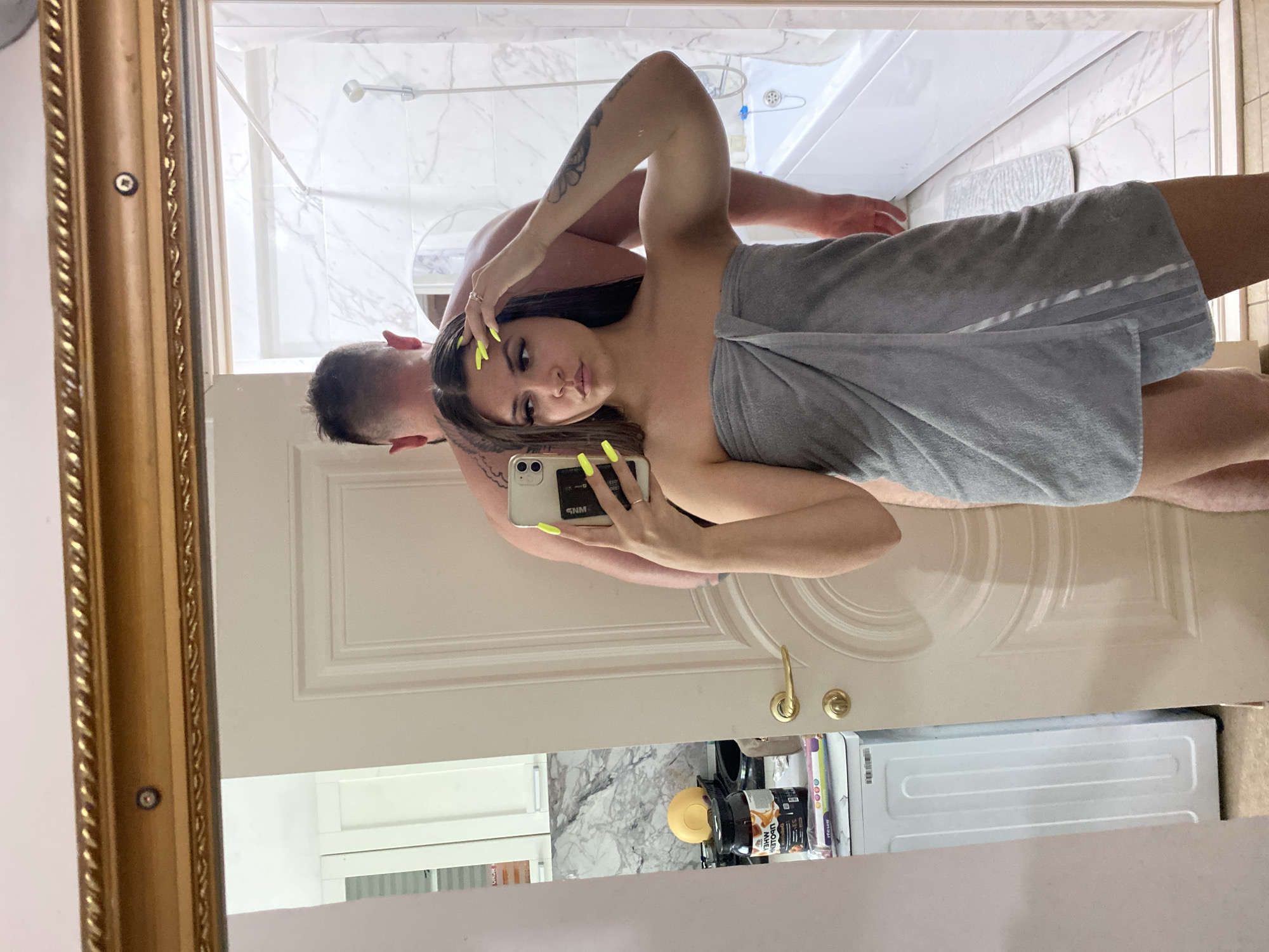 A woman in a grey towel is taking a mirror selfie, with a man standing behind her. The woman is in the foreground, facing the mirror, with her right arm raised to hold the phone. She has long dark hair, dramatic eye makeup, and bright yellow acrylic nails. She is wearing a grey towel wrapped around her body. Behind her, a man is visible, also partially obscured by the woman. His back is to the camera, and he has tattoos on his arms and back. They are standing in a bathroom, as evidenced by the bathtub and tiled walls visible in the reflection. The room appears to be in a residential setting. The image also shows a doorway and a kitchen area with a washing machine and some products on a counter. The overall lighting is bright, suggesting the photo was taken during the day or with good artificial lighting. The setting does not provide any specific clues that would place it in Makhachkala, Russia.