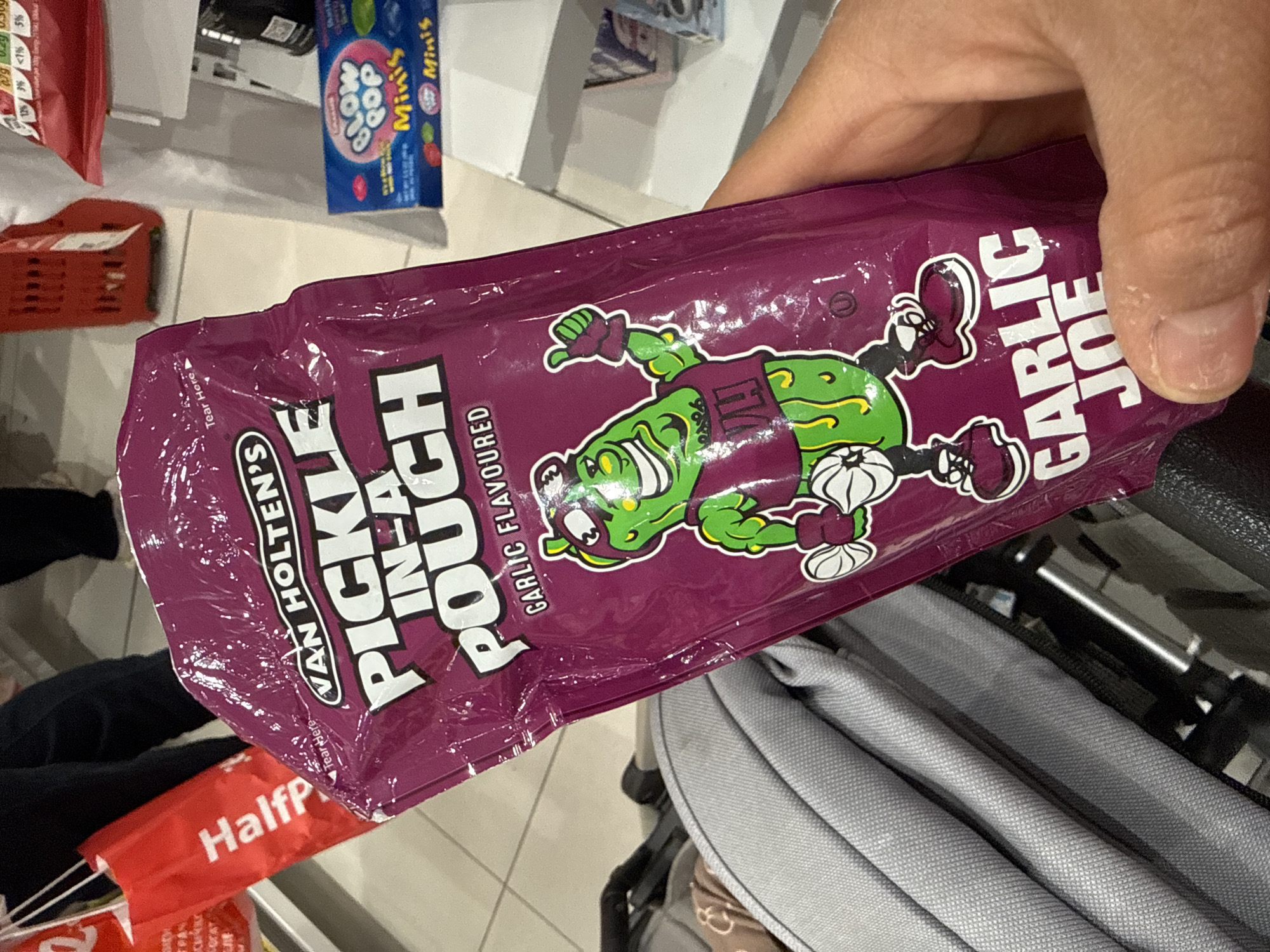 The image shows a purple "Van Holten's Pickle-in-a-Pouch Garlic Flavored" package being held in a person's hand. The bag features a cartoon pickle character wearing a shirt and cleats. The background shows other items in what appears to be a store or shopping environment. A red and white "Halfp" sign is visible, and a blue box of "Blow Pop Minis" can be seen on a shelf in the background. The bottom right corner of the image shows a grey stroller or buggy. There is no information present to determine the city or country in which this photograph was taken.