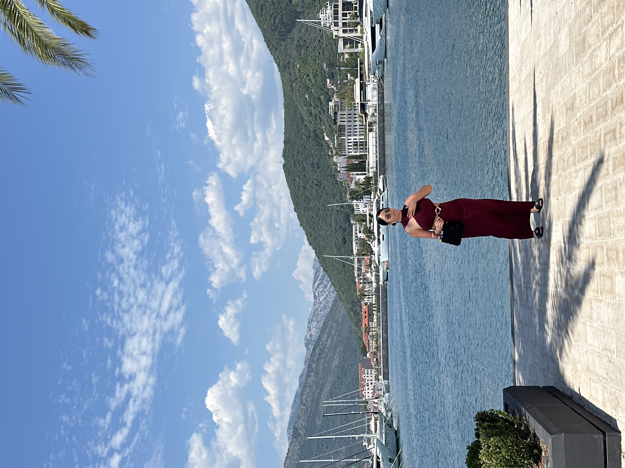 A woman stands on a paved walkway beside a body of water in Donja Lastva, Montenegro. She is wearing a long, maroon dress with a halter neckline and a gold belt. She is holding a black purse in her left hand and has her right hand on her chest. She wears sunglasses. In the background, several white sailboats are moored in the water. Beyond the boats, a town with red-tiled roofs is visible, nestled at the base of lush green mountains. The sky is blue with scattered white clouds. A palm frond is visible in the upper right corner of the image. The walkway is made of light-colored, rectangular paving stones, and a dark planter with green bushes is to the left of the woman. The sun casts shadows on the walkway.