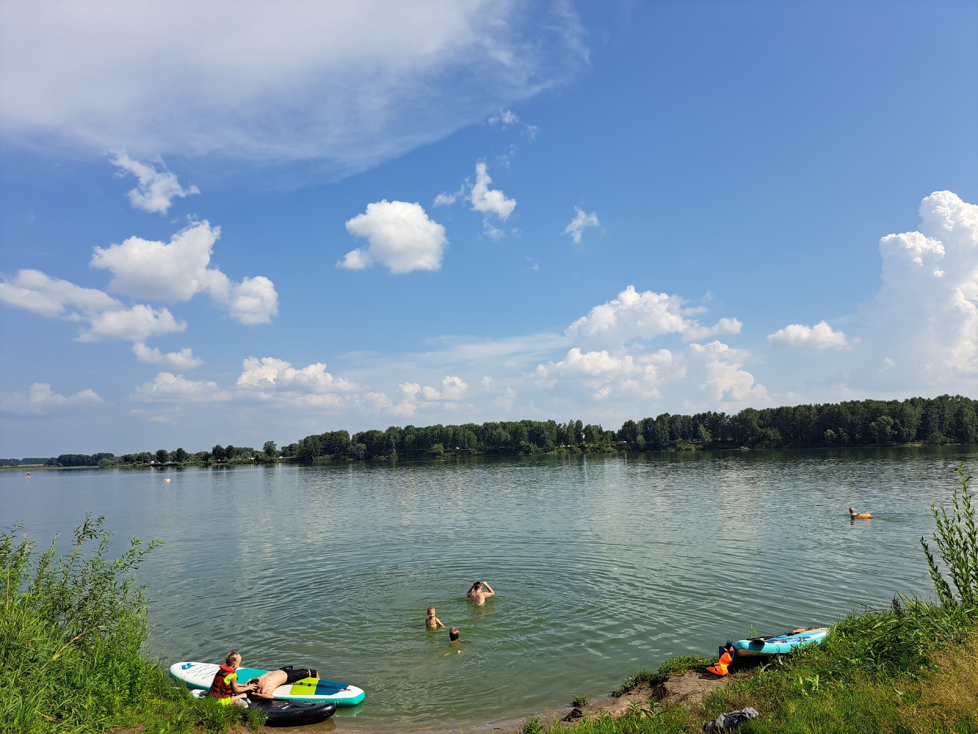 The image depicts a serene lake scene on a bright, partly cloudy day. In the foreground, on the grassy bank, are two paddleboards and a life vest. To the left, a person in a life vest is lying on a paddleboard in the shallow water, while another person lies on a separate paddleboard next to them. Further out in the lake, three individuals are swimming. In the distance, on the far side of the lake, a dense line of trees forms the horizon, with a few scattered buildings visible amongst them. A person is visible in the water on the right side of the lake, appearing to be using a floating ring. The lake water is calm with gentle ripples. The sky is a vibrant blue with scattered white cumulus clouds. There are no specific landmarks or identifiers to pinpoint the location within the city of Sorochii Log, Russia.