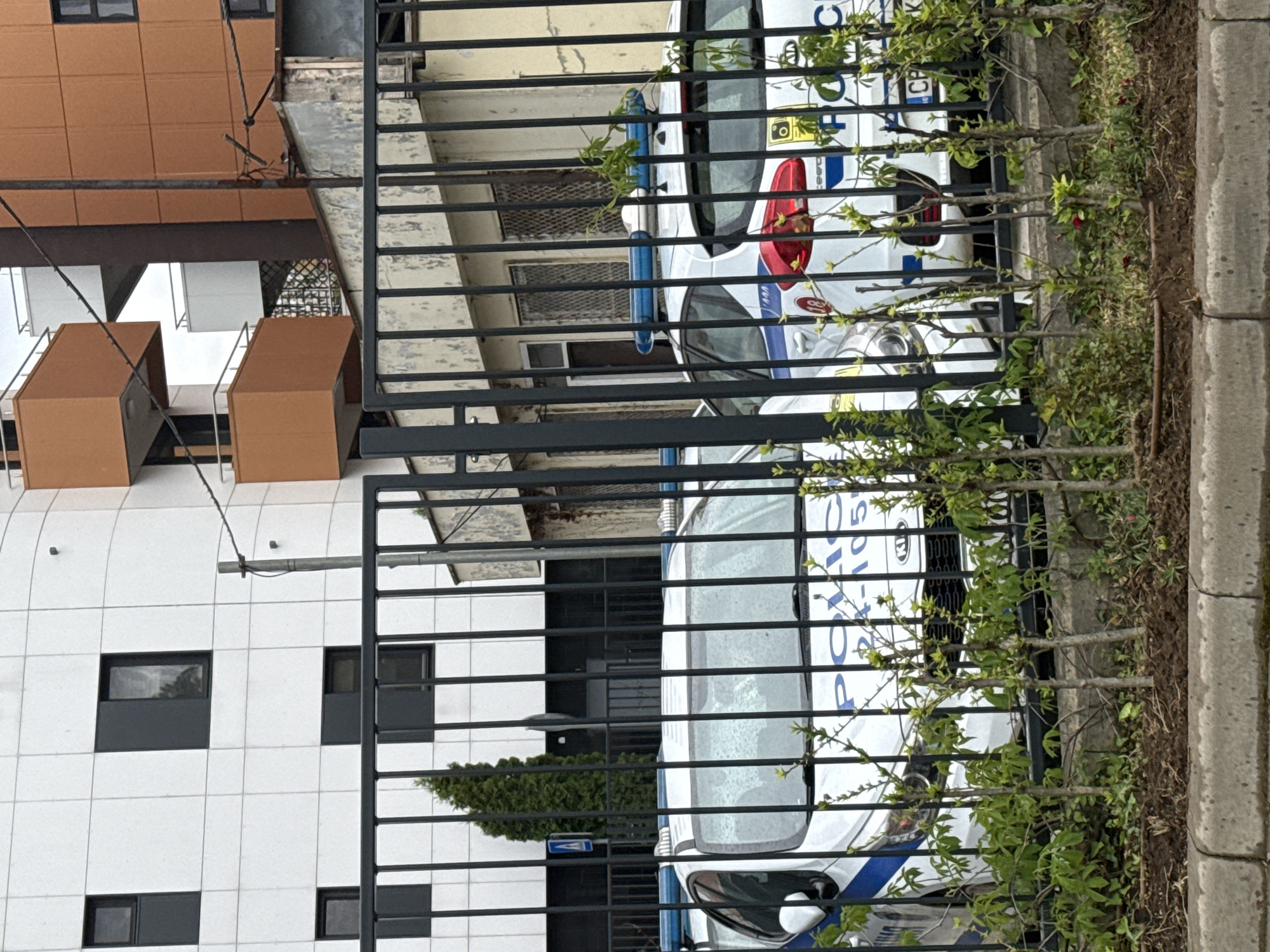The image shows two Bulgarian police cars parked behind a black metal fence next to a modern building. The building has a white facade with dark-framed windows, and sections with brown cladding. The foreground contains vegetation growing near the fence. Based on the visible lettering "ПОЛИЦИЯ" (Police) on the vehicles, they are Bulgarian police cars. Based on the "CP" license plate suffix, it is somewhere in Sofia, Bulgaria. The scene is likely in an urban area, given the presence of modern architecture and fenced-off police vehicles. The image depicts a static scene with no visible people or actions. The presence of police vehicles suggests a security presence or potential activity in the area. The weather conditions are unknown.