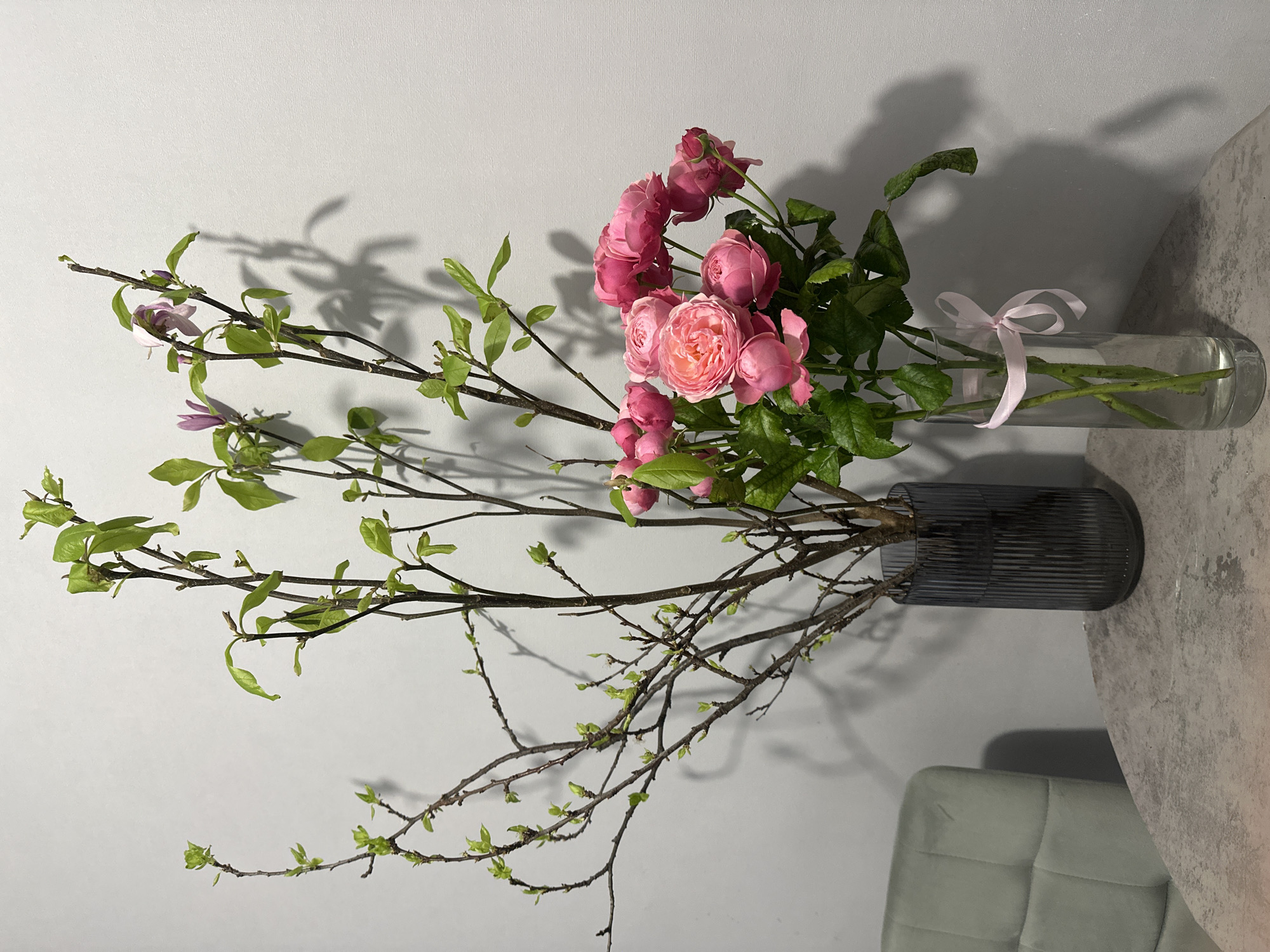 The image shows a floral arrangement with two vases. One vase is dark gray and ribbed, containing bare branches with nascent green leaves, and some branches with larger green leaves and a few purple flowers. The other vase is clear glass and contains roses with green leaves and stems, tied with a pink ribbon. The arrangement is placed on a gray surface against a light gray wall. A section of a gray cushioned chair is visible in the bottom right corner. The image appears to be indoors, possibly in a home or studio. There is no information to suggest the photo was taken in Kazan, Russia.