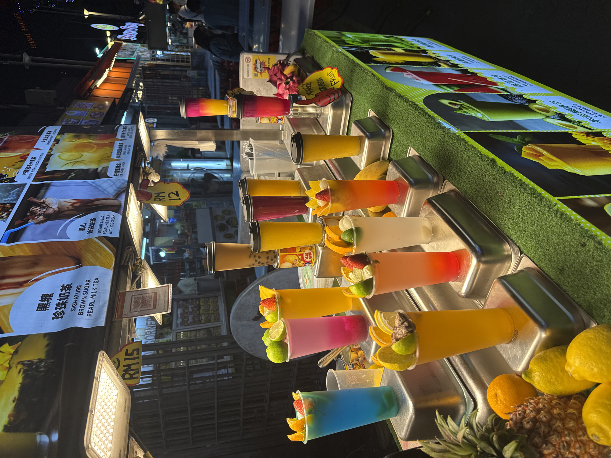This image depicts a vibrant night market stall, likely in Kuala Lumpur, Malaysia, illuminated by artificial lights, indicating it is nighttime. The stall specializes in colorful fruit beverages and pearl milk tea, displayed prominently in transparent cups arranged in metal holders on a counter.
A wide variety of drinks are shown, exhibiting bright hues of red, orange, yellow, pink, blue, and brown. Many are garnished with fresh fruit slices like lemons, limes, oranges, and strawberries, enhancing their appeal. Some cups contain tapioca pearls, typical of bubble tea. Fresh fruits such as pineapples, lemons, and oranges are visible on the counter, suggesting their use in the drinks. The display also features a green artificial turf surface with large pictorial menus of various fruit juices and smoothies, including "AVOCADO" and "DRAGON FRUIT."
In the background, other market stalls and blurred figures of people suggest a busy, lively atmosphere. Visible text on the display boards advertises specific drinks and their prices. Drink options include "SIGNATURE BROWN SUGAR PEARL MILK TEA," "BROWN SUGAR PEARL MILK TEA WITH ICE CREAM," "LEMON WITH PASSION FRUIT TEA," "DRAGON FRUIT LEMONADE," "ICE HONEY LEMON," "FRESH ORANGE," and "KIWI FRUIT TEA." Prices like "RM 15" and "RM 12" are clearly marked in Malaysian Ringgit. Other text includes "NO SMOKING," "FRUIT," and the names "IZUAL HOTEL" and "Solay" on signs in the distant background. The scene conveys a bustling and appealing street food vendor environment.