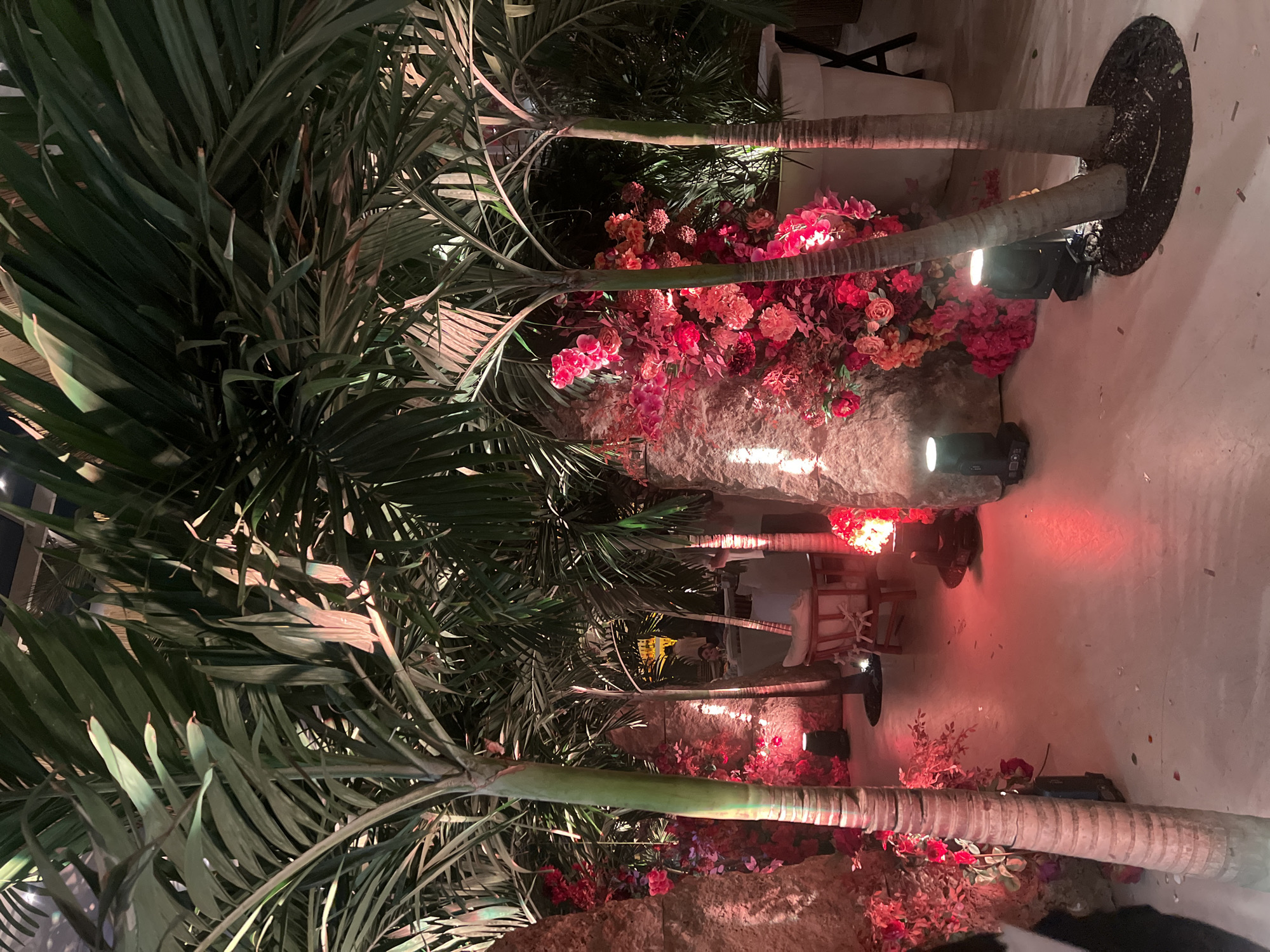 The image shows a landscaped indoor space, likely a venue for an event. There are palm trees arranged in a row, interspersed with floral arrangements featuring red and pink flowers. Spotlights illuminate the scene with red and white light. Chairs and a table are visible in the background, suggesting a seating area. The floor is a light-colored concrete or similar material. The location is possibly Yerevan, Armenia, but there's no specific visual evidence to confirm it within the image. There are no people clearly visible.