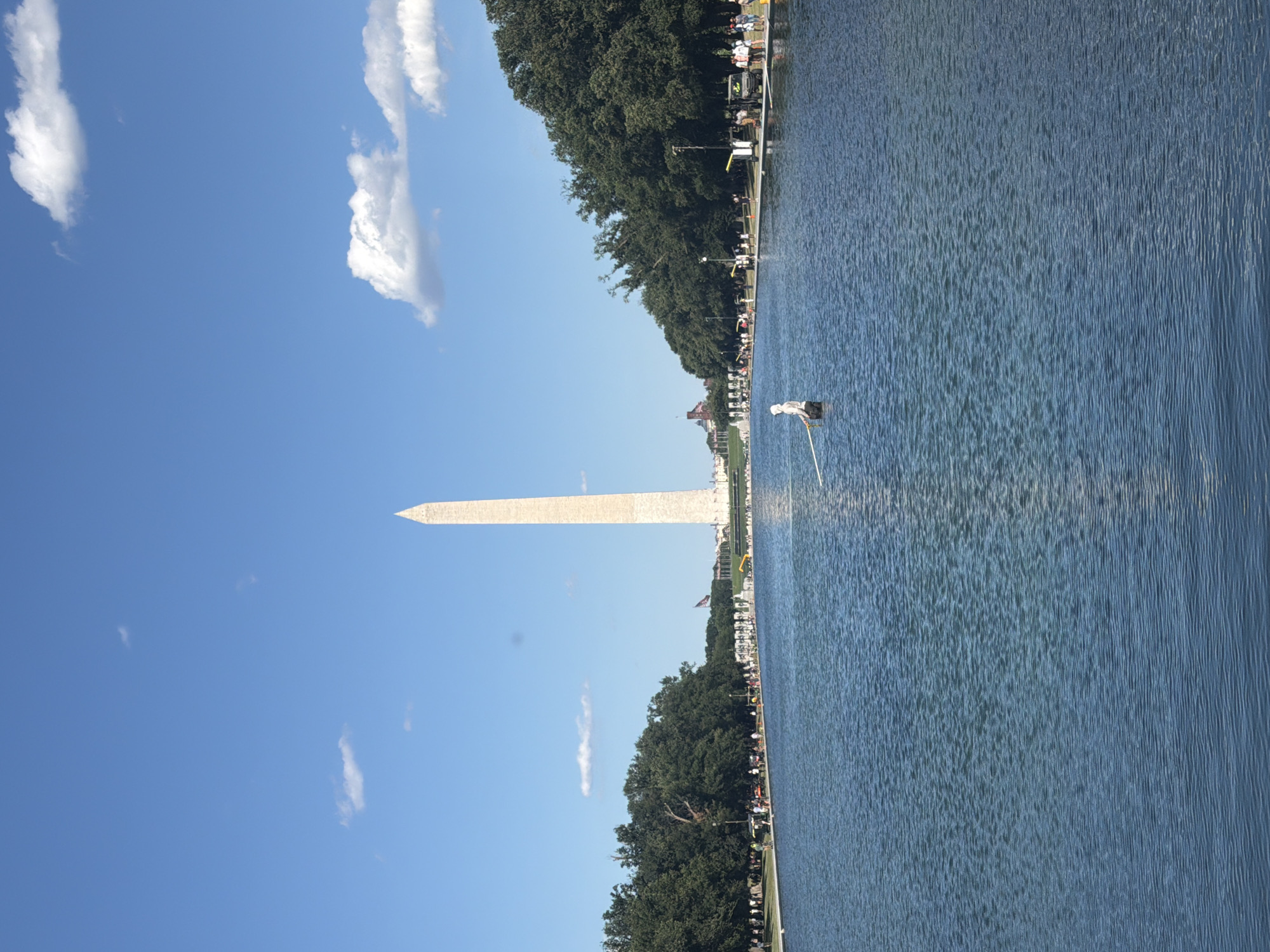 The image depicts the Washington Monument in Washington, D.C., viewed from across the Reflecting Pool. The sky is a clear blue with scattered white clouds. The Washington Monument, a tall, slender obelisk, stands prominently in the distance. In the foreground, the Reflecting Pool occupies the majority of the frame, with gentle ripples on its surface. A lone figure, seemingly wading in the shallow edge of the pool, is visible in the middle ground, holding a long pole. Along the far edge of the pool, a line of trees borders the scene, with people and vehicles present near the tree line. The overall scene suggests a sunny day in a public space within the city.