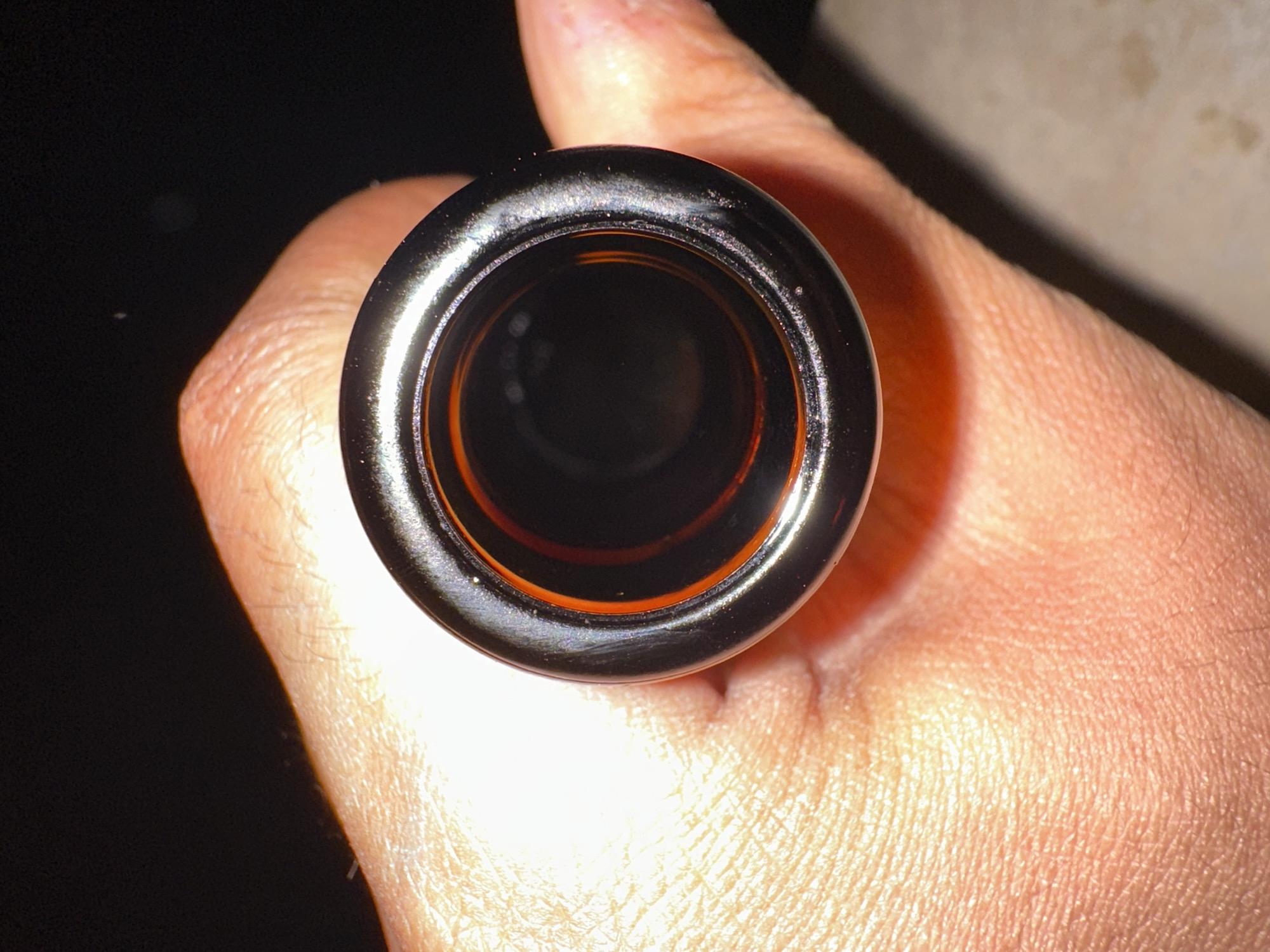 The image shows a close-up, overhead view of a person's finger and thumb holding a cylindrical object. The object appears to be metallic with a dark, shiny exterior, and an orange interior. The person's skin texture is visible, with fine lines and hairs. The background is dark, with some out-of-focus lighter areas suggesting a dimly lit environment. The lighting is focused on the object and the hand, creating highlights and shadows. The overall impression is one of a detailed, macro-style shot of everyday objects. No text is visible in the image. The location context provided is Dilijan, Armenia, but this information cannot be visually confirmed from the image itself.