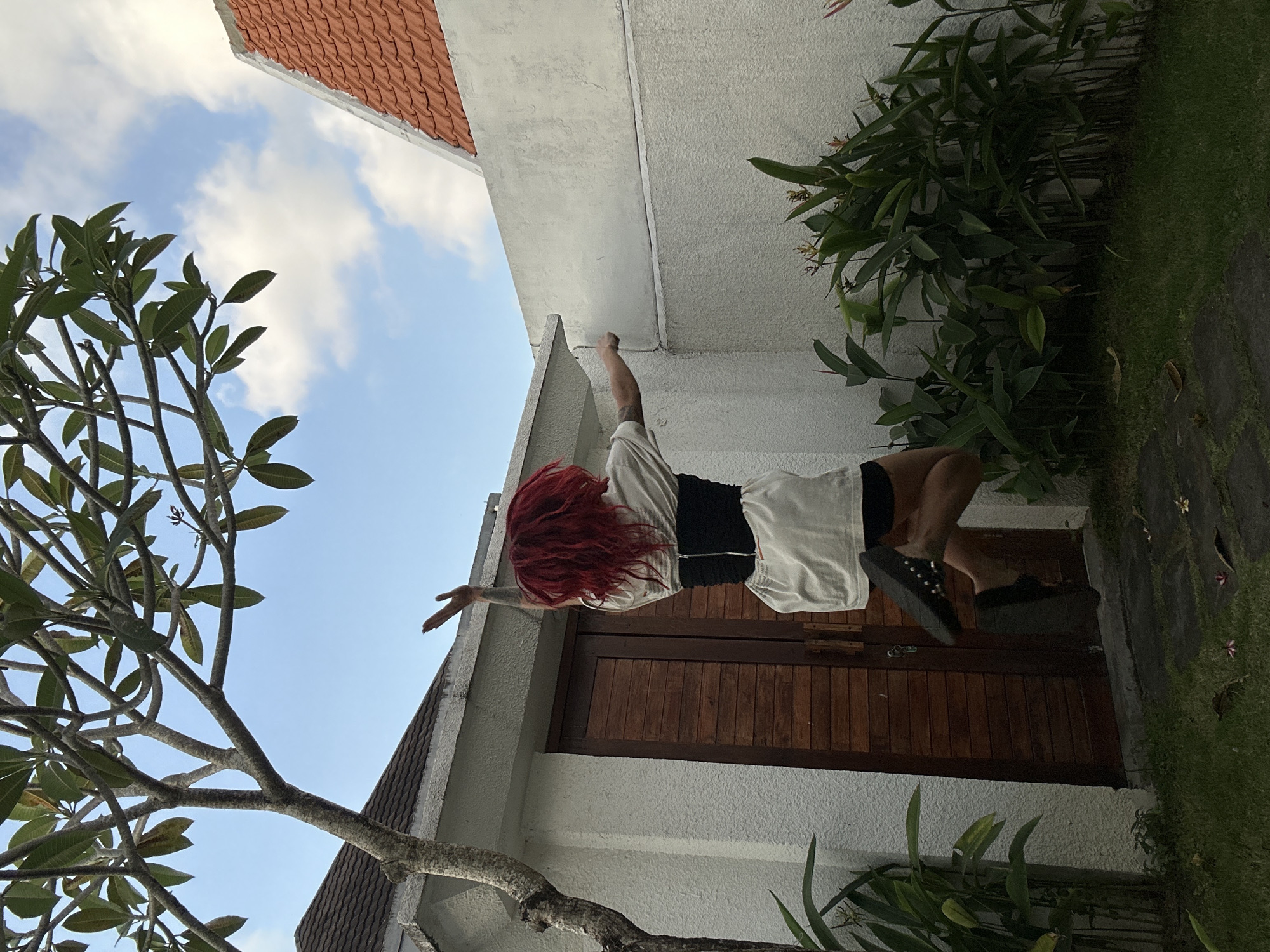 The image depicts an outdoor scene in North Kuta, Indonesia, featuring a person in mid-jump within a private garden or courtyard. The individual, seen from the back, has bright red, textured hair and is wearing a white oversized t-shirt, black shorts, and a black corset-style waist cincher over the t-shirt. Black platform shoes are visible on their feet. Both arms are raised and bent, with hands open and fingers spread; a tattoo is visible on the left arm. The person is suspended approximately 0.5 to 1 meter above a grass patch, with bent knees and feet off the ground, indicating an energetic leap.
The environment includes a prominent tree with light-colored, bare branches and green leaves, possibly a Plumeria, positioned in the upper left of the frame. Below and to the left of the person is a white stucco building featuring a double wooden door with horizontal paneling and dark hardware. Above the door, a light brown tiled roof is partially visible, along with white concrete eaves. To the right, a white stucco wall extends, meeting another wall at an angle, with a prominent horizontal line across its surface. Beyond this wall, the orange-red terracotta tiled roof of another structure is visible against a clear blue sky with scattered white clouds. The ground consists of short green grass, interspersed with irregular dark gray stone pavers in the foreground. Dense green tropical foliage and bushes line the base of the white walls, with some plants exhibiting slender stalks and hints of reddish-orange blossoms. Scattered dry leaves and small pinkish flower petals are present on the grass. The lighting suggests daytime with soft, natural illumination.