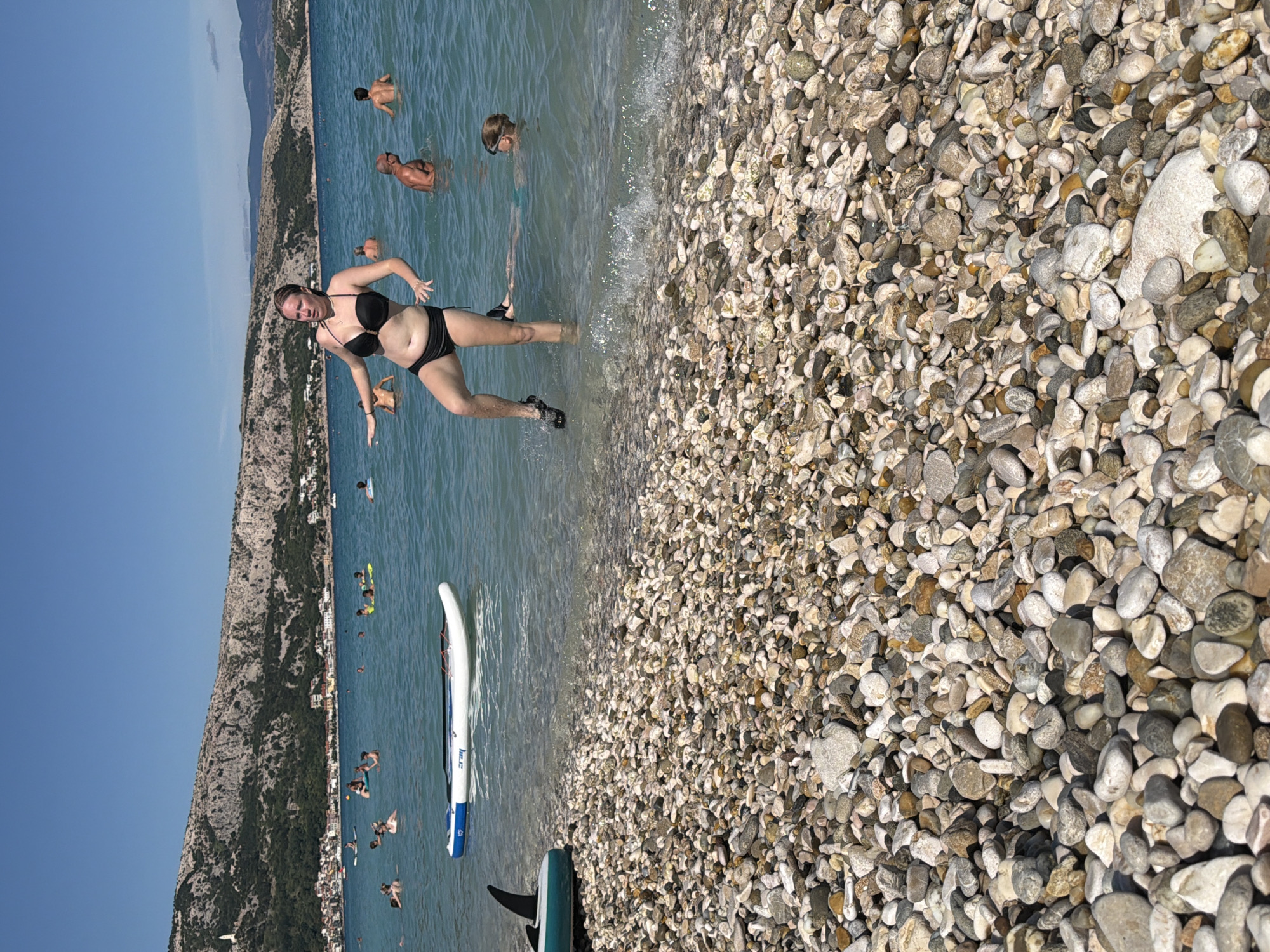 The image depicts a crowded pebble beach with clear blue water, a rocky hillside dotted with buildings in the background, and a bright blue sky. In the foreground, the beach is covered with a variety of smooth, multi-colored pebbles. Several people are in the water. One woman in a black bikini stands in the shallow water, with her right leg raised and her left arm extended. Other individuals are seen swimming or wading further out. A white and blue paddleboard floats on the water near the shore. To the left, the fin of a teal-colored board is visible. The location is identified as Općina Baška, Croatia, characterized by its coastal environment and the presence of a town nestled against a mountainous backdrop. The overall scene is one of a sunny, active day at the beach.