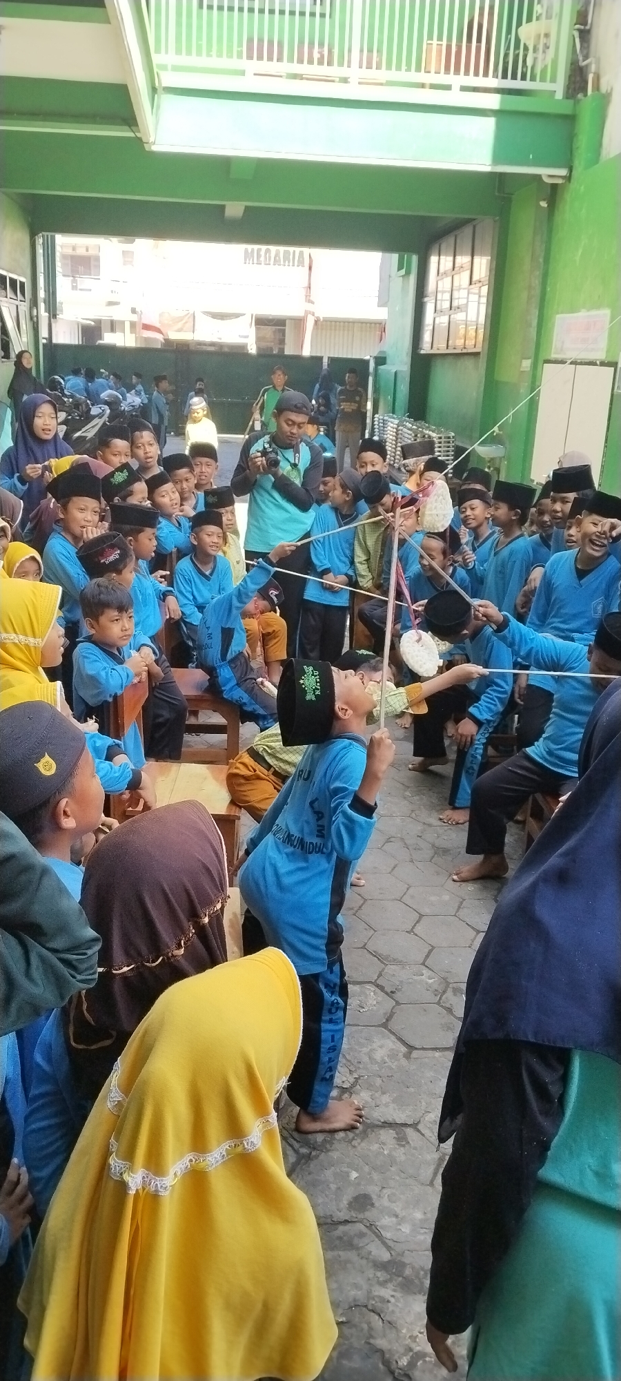In Yosowilangun, Indonesia, a group of children are participating in a cracker-eating game, a common Ramadan tradition. The children, mostly boys, are dressed in blue shirts and black peci hats. They stand in a line, each holding a string with a cracker attached, attempting to eat it without using their hands. Some are focused, their faces tilted upwards, while others are laughing and cheering. In the background, a man is capturing the event with a camera. The setting appears to be a school or community center, with green walls and a white railing visible. The ground is paved with stone tiles. The overall atmosphere is one of lively celebration and community.