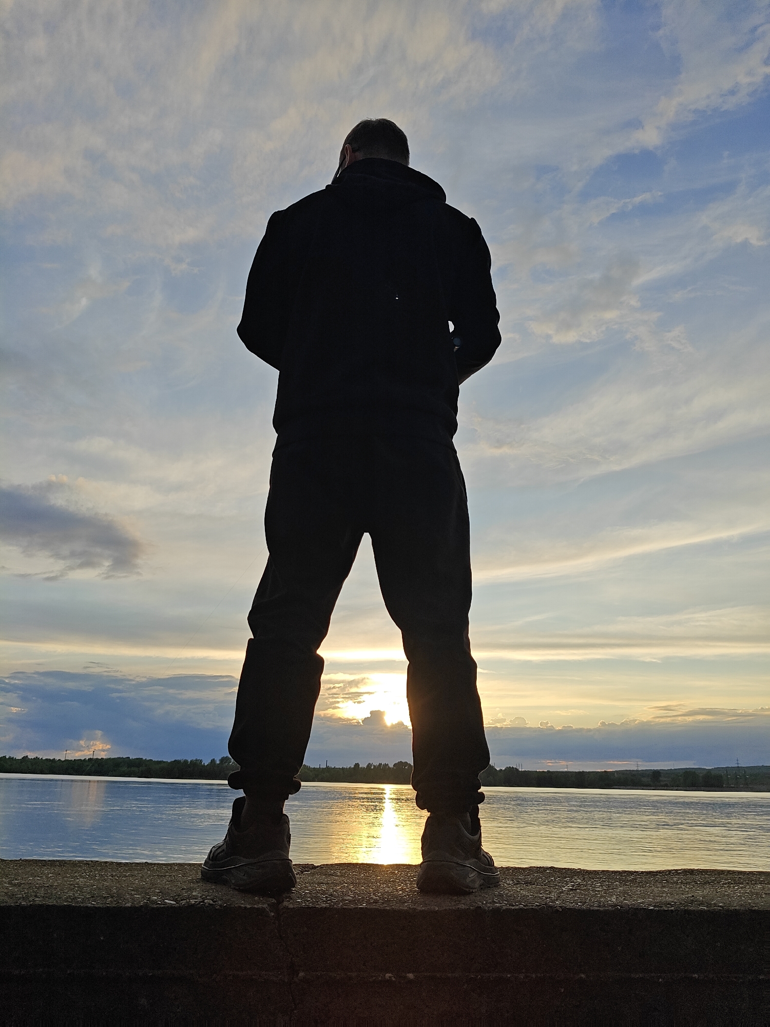 A person stands with their back to the viewer, facing a body of water at sunset. The individual is wearing a dark hooded sweatshirt and dark pants. They are standing on a concrete structure, possibly a dam or embankment. The setting sun is reflecting off the water, creating a bright glare. The sky is filled with scattered clouds. In the distance, beyond the water, there is a line of trees and some buildings, possibly indicating the opposite shore of the water body. The location is likely Chaikovsky, Russia, given the prompt's context.