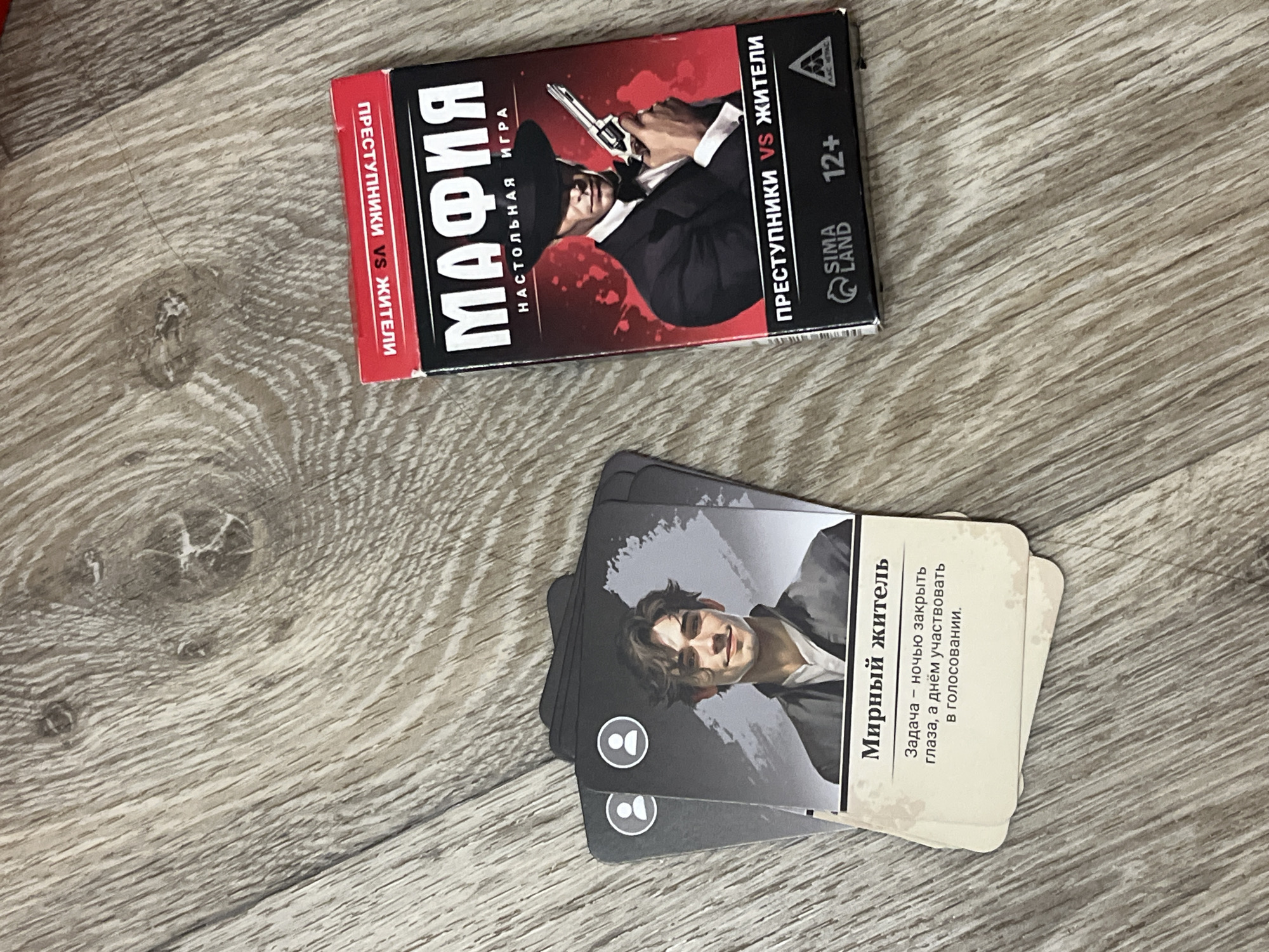 The image shows a board game called "Mafia: Desktop Game" placed on a wooden surface. The game box is predominantly red and black, featuring an illustration of a man in a suit and hat holding a gun. The cards of the game are fanned out in front of the box, with one card clearly visible showing a portrait of a young man and text in Russian, identifying him as a "Peaceful Resident" and describing his role in the game. The wooden surface appears to be a floor with a visible grain and some knot patterns. The lighting suggests an indoor setting.