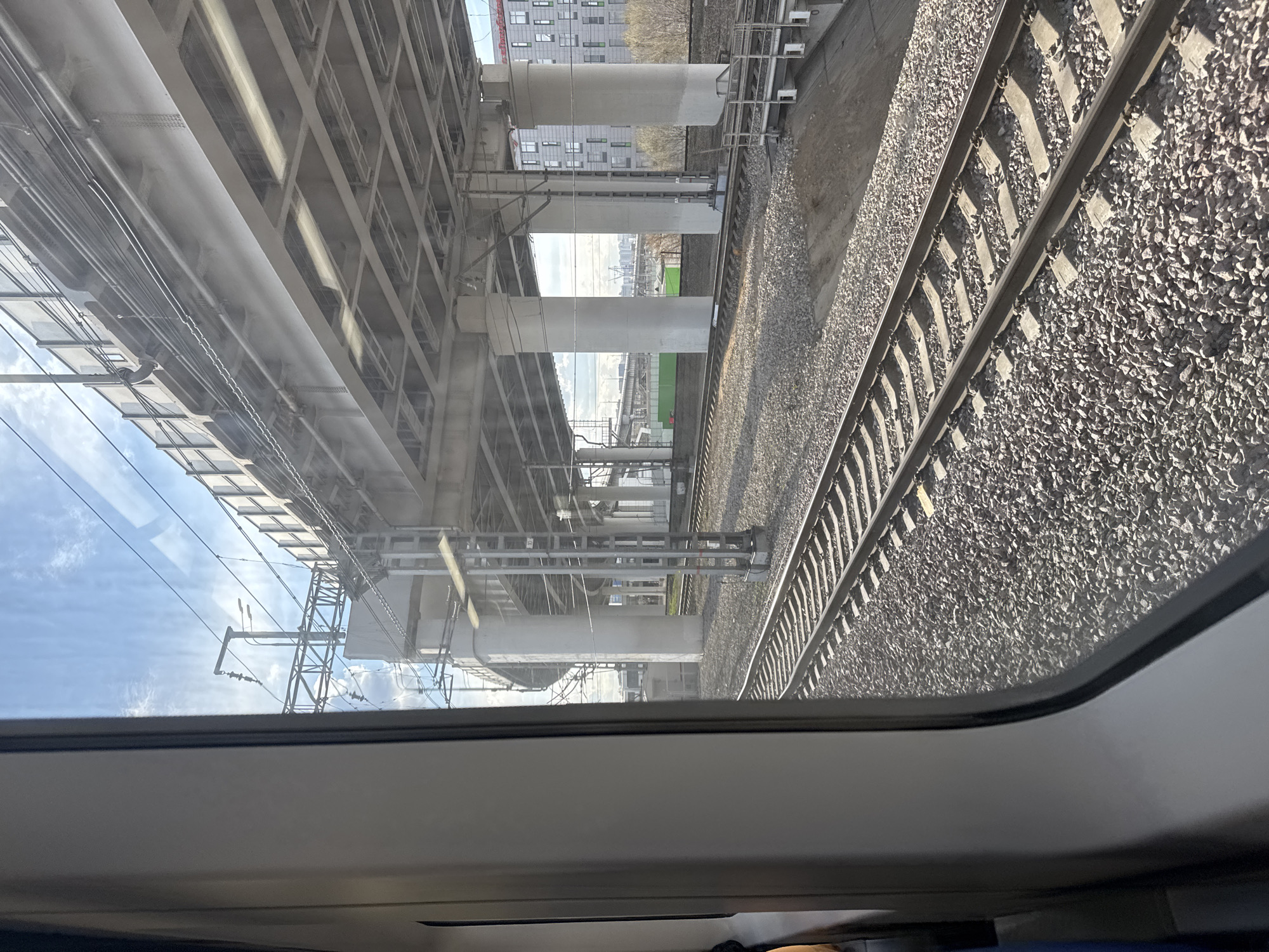 The image shows a view from inside a train, likely taken in Khimki, Russia, as suggested by the presence of typical urban infrastructure and building styles in the background.
The scene is dominated by two parallel railway tracks filled with grey gravel. Adjacent to the tracks is a large, elevated concrete structure, likely a bridge or an overpass, supported by thick cylindrical pillars. Overhead, the sky is visible through the window, partly cloudy with a hint of blue. Reflected on the window is the blue sky.