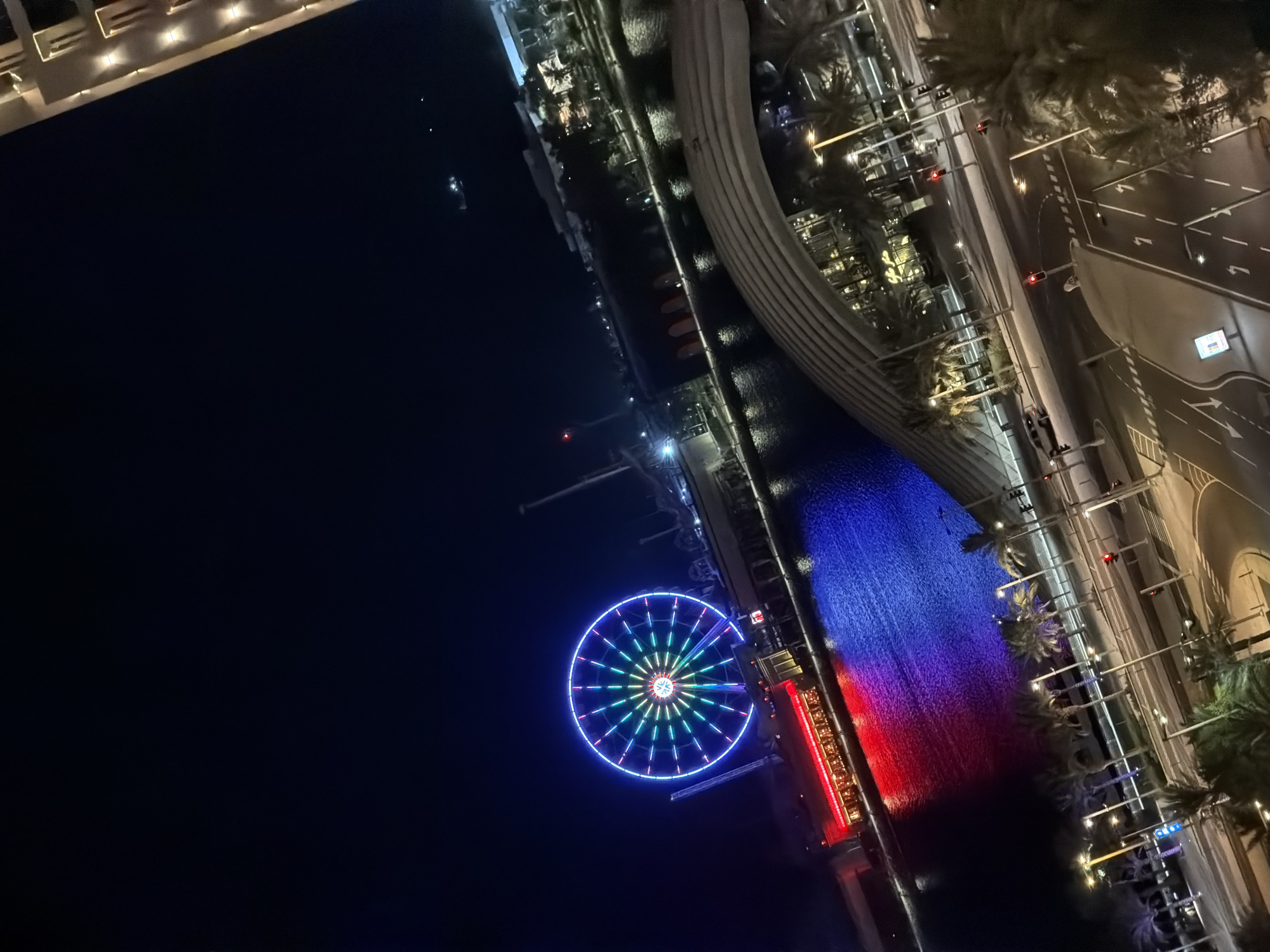 The image captures a nighttime view of a waterfront area in an unknown city. A large, brightly lit Ferris wheel dominates the scene on the left, displaying a vibrant spectrum of colors including blue, green, and pink. Adjacent to the Ferris wheel is a building illuminated with red lights. The water in front of these structures is a deep blue, reflecting the colorful lights from the Ferris wheel and the building, creating a dramatic effect of red and blue illumination on the water's surface. In the background, more city lights and structures are visible, along with palm trees lining a waterfront promenade. A curved, modern architectural structure with lighting is prominent in the lower right quadrant of the image. Below this structure, a street with traffic lights, road markings, and a few parked cars is visible. The overall atmosphere is one of a lively, illuminated urban entertainment district at night.