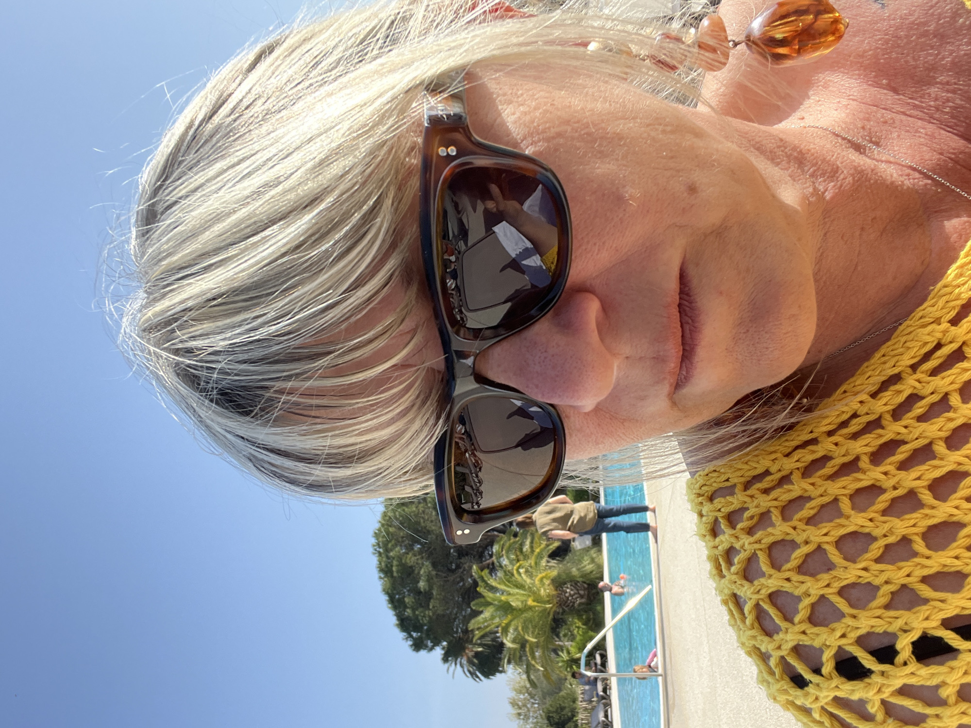 A woman with sunglasses and fair hair stands in the vicinity of a swimming pool in Grimaud, France. The pool is visible in the reflection of her sunglasses and the lower portion of the frame. There are people in and around the pool. Palm trees and other greenery are also visible in the background. The woman is wearing a yellow crocheted top and a necklace with a large amber-colored bead.