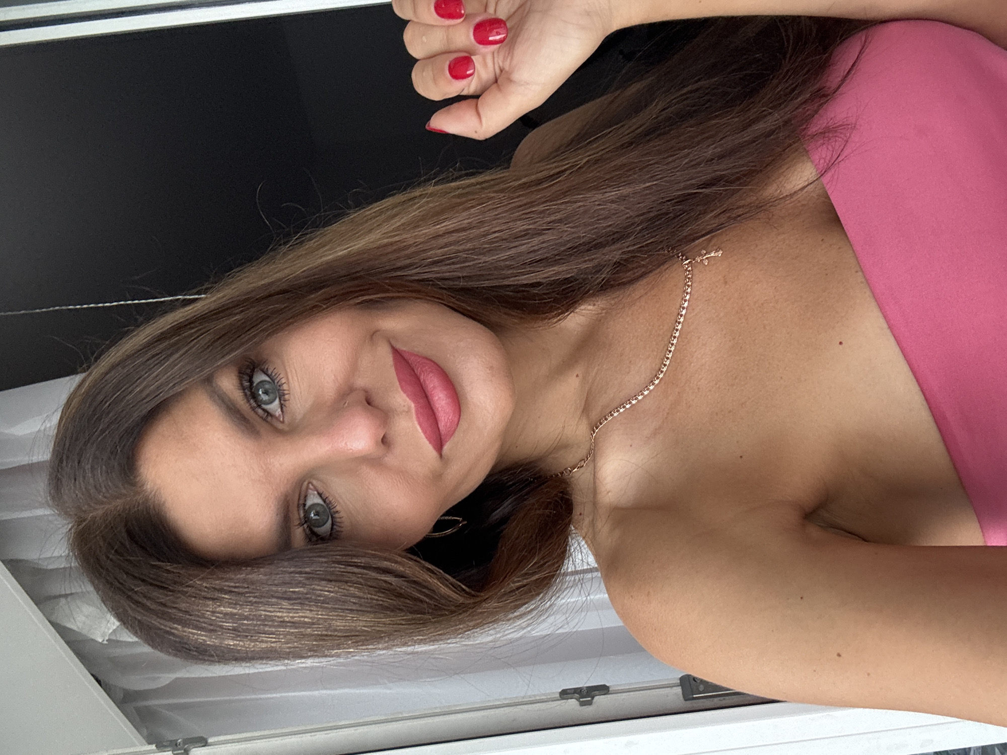 A woman with long brown hair and blue eyes, wearing a pink off-the-shoulder top and a gold necklace, poses for a selfie. She has bright red nail polish on her fingernails, and she is wearing subtle makeup. Her right hand is visible, raised in front of her. The background is a mix of a white curtain and a dark wall. The image does not depict Chaikovsky, Russia.