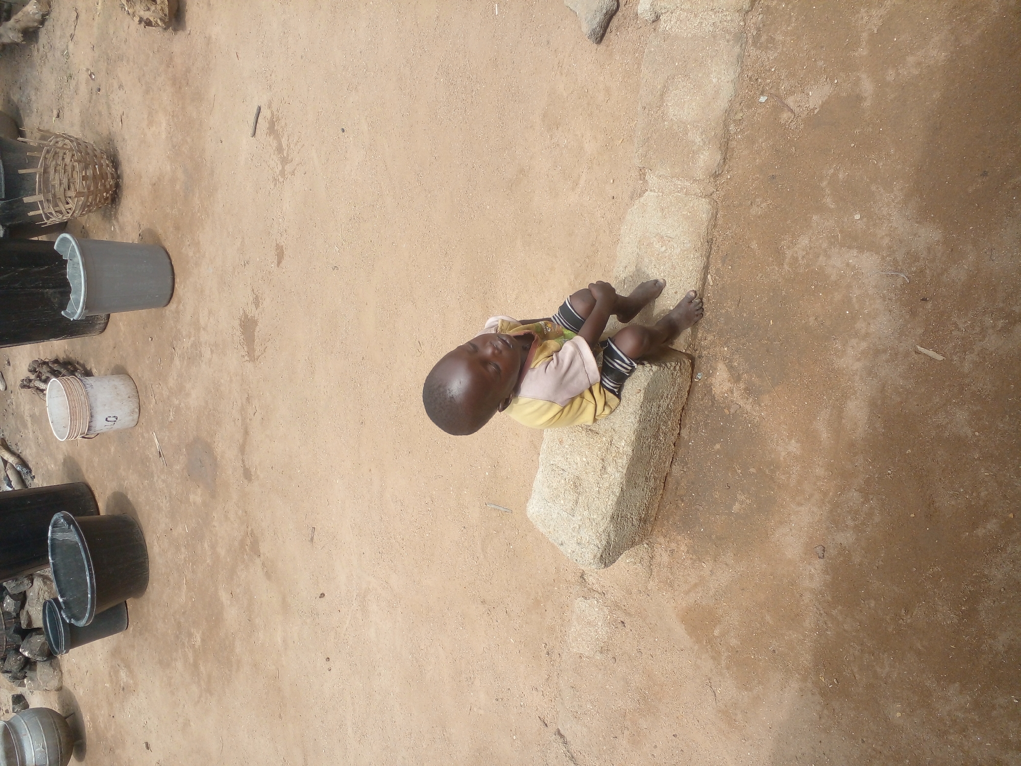 A young boy sits on a large rock in a dusty, outdoor area. He is wearing a yellow t-shirt with a design and black and white striped shorts. His arms are resting on his knees, and his head is tilted down, with his eyes closed. The ground is a mixture of sand and dirt, with a few scattered pebbles and dried leaves. In the background, there are several large, dark-colored buckets and a woven basket. The overall impression is one of a simple, rural setting.