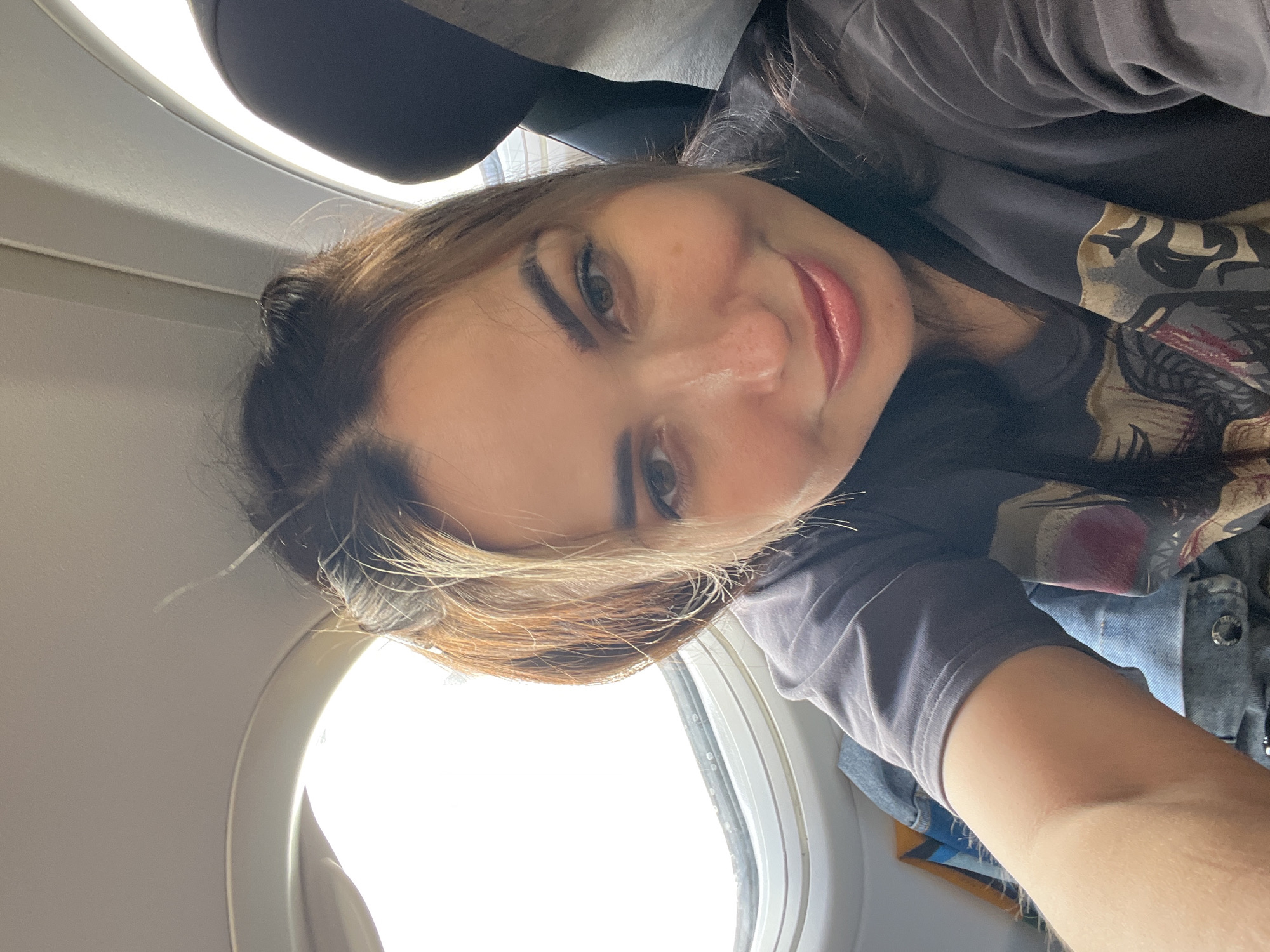 A woman is taking a selfie on an airplane. She is wearing a dark gray t-shirt and has her hair pulled back in two buns. The window of the airplane is to her left, and a headrest is visible to her right. She is smiling and looking at the camera. The lighting is bright, likely from the sun outside the window.