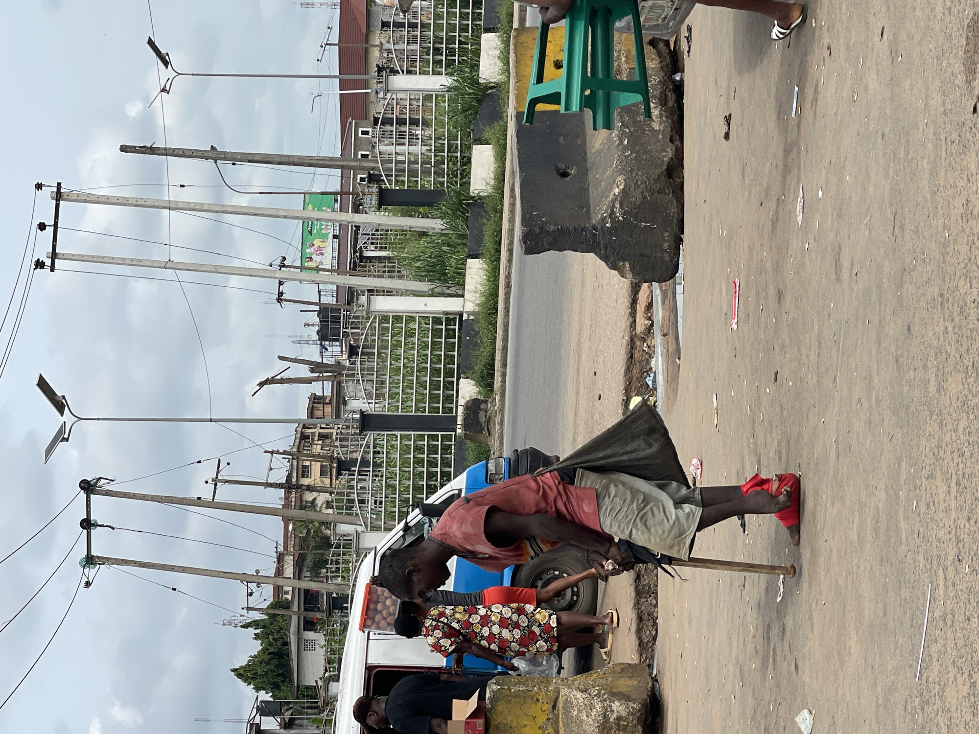 The image depicts a street scene in Onitsha, Nigeria. A man with a physical disability uses a crutch for support while standing on the side of a road. He is wearing a red shirt, grey shorts, and red footwear. He is interacting with individuals standing near a blue and white minivan, possibly making a purchase. Other people are present near the vehicle, engaging in what appears to be informal commerce. The background features utility poles with electrical wires, buildings, and vegetation. The roadside shows evidence of wear and litter. The scene suggests a typical day in an urban, commercial environment.