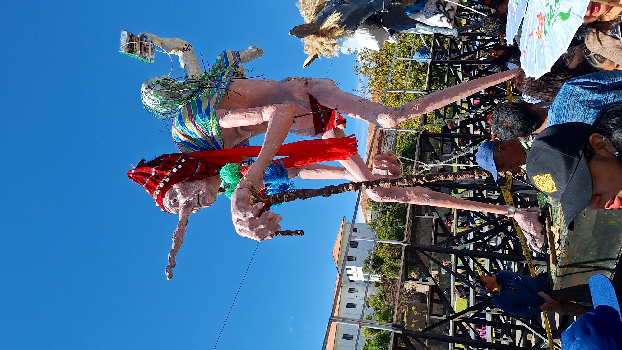 This image captures a vibrant festival scene in Cuzco, Peru, likely during a parade or procession. The focal point is a colossal effigy of a person with exaggerated features, including a long, prominent nose and thin, elongated limbs. The effigy is adorned with colorful traditional attire: a red and white striped hat, a bright red scarf, and a striped sash across its chest. It holds a gnarled cane in its right hand, decorated with blue and green pom-poms. A smaller figure, also appearing to be part of the effigy or a prop, is positioned on its back, holding a device resembling a smartphone or small tablet. The effigy stands on a platform, supported by a dark, scaffolding-like structure, and is surrounded by onlookers.
In the background, white buildings with red-tiled roofs, typical of colonial architecture, line a street. Green trees and foliage are visible, suggesting an outdoor urban setting. Several people are present in the foreground, some wearing casual clothing, while others are partially obscured by the effigy. A large, gray structure with a horse's head and mane is visible to the right of the main effigy, indicating the presence of other parade floats. Yellow caution tape with "PELIGRO NO PASAR" (Danger Do Not Pass) is visible along the scaffolding, a common sight at public events to manage crowds and ensure safety. The sky is a clear, bright blue, signifying a sunny day. The overall atmosphere suggests a lively cultural celebration.