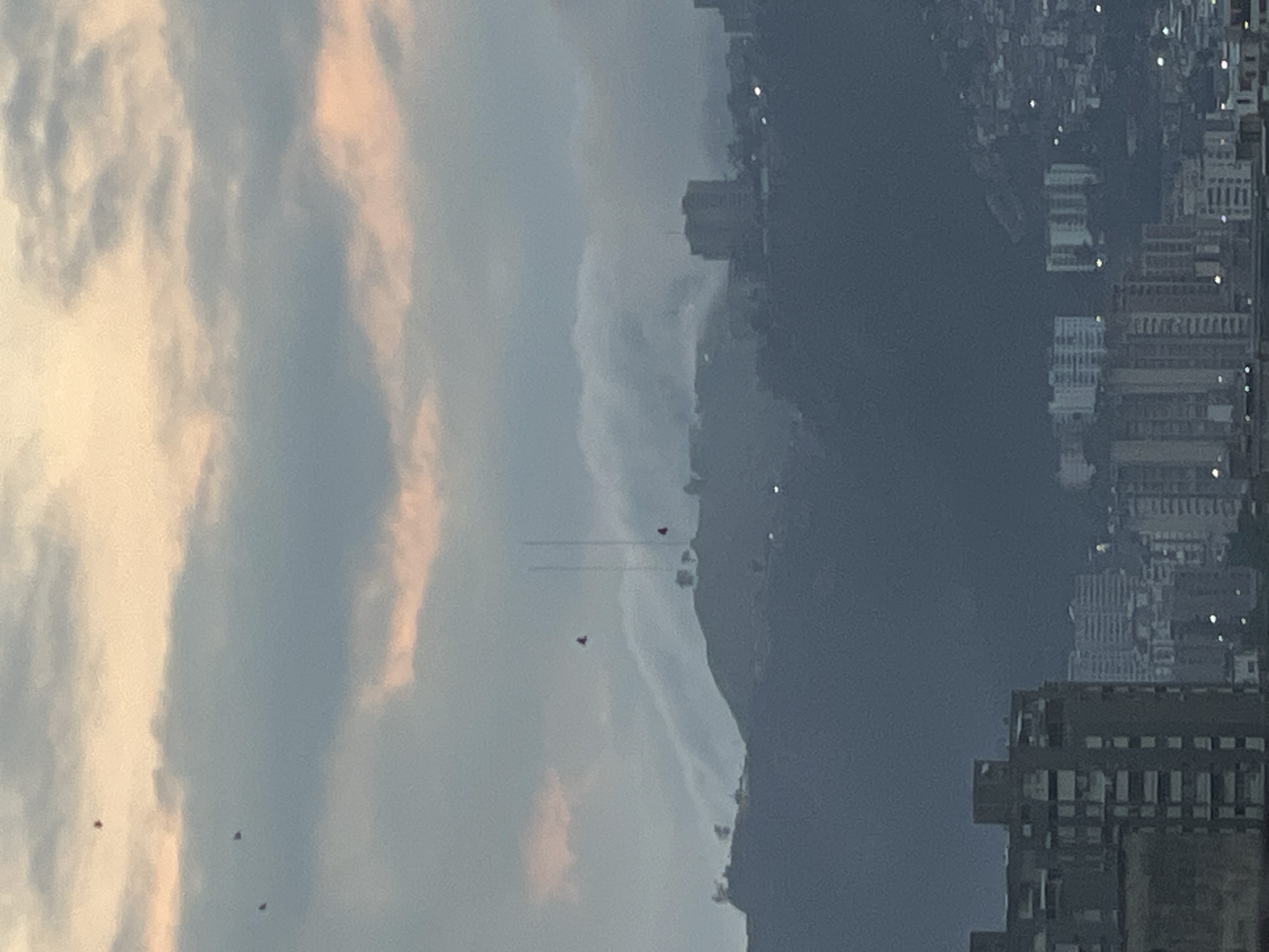 The image shows a cityscape in Caracas, Venezuela, with a sky dominated by clouds. The building structures are dense and illuminated by dispersed light. The clouds are scattered with tinges of color. There are a few birds in flight within the view.