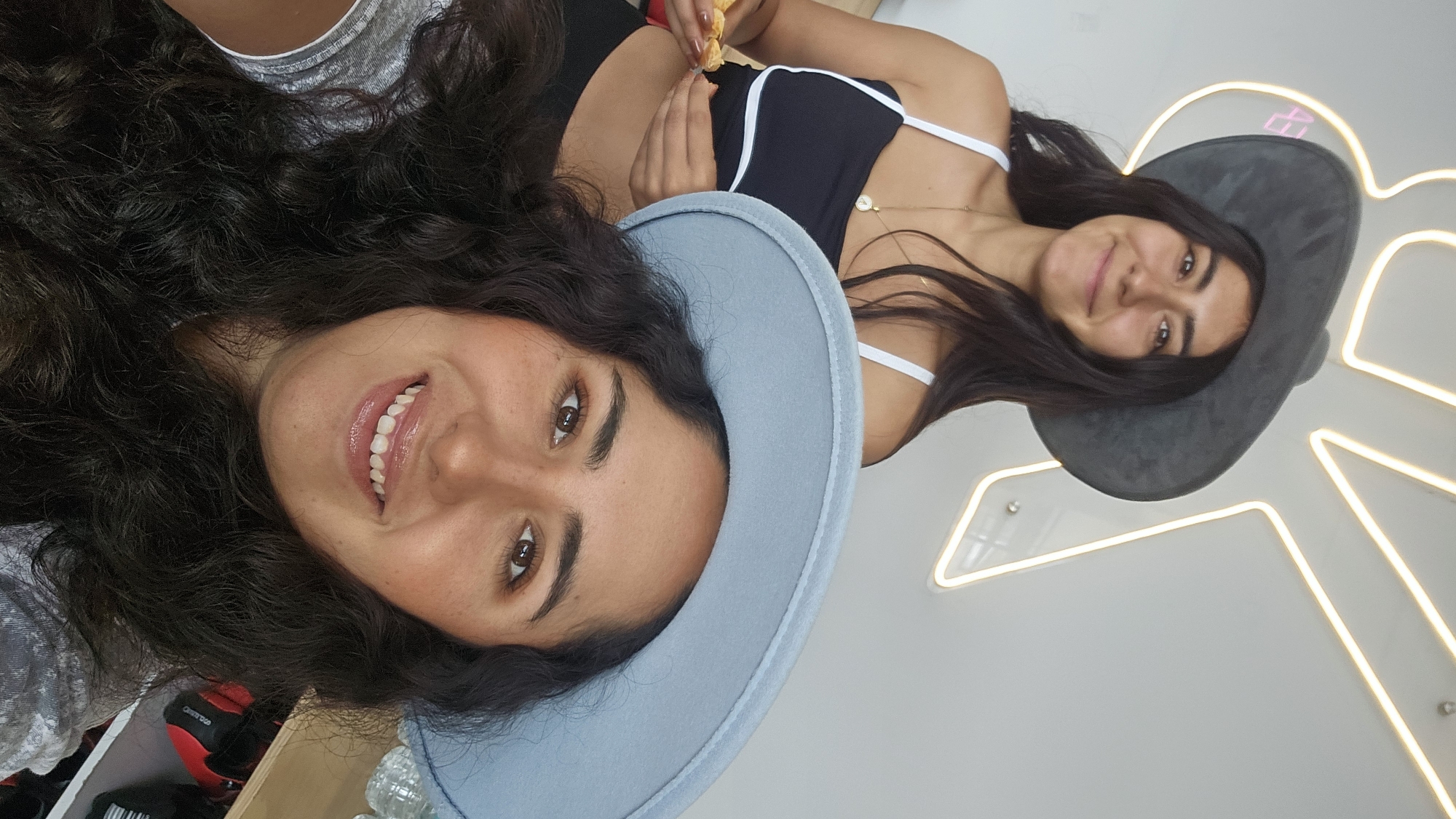 An indoor image captures two young women, oriented 90 degrees clockwise, smiling brightly. The woman in the foreground is in a close-up, her head and shoulders filling the left side of the frame. She has long, dark, curly hair, brown eyes, and is wearing a light blue, felt-like hat. Her lips are parted in a smile, revealing white teeth, and her cheeks show a touch of blush. She wears a light-colored top, partially visible.
Behind her, slightly out of focus, is another young woman. She has straight, dark hair and a darker, perhaps charcoal-grey, felt-like hat. She is wearing a dark top with thin white straps and a delicate gold necklace with a circular pendant. Her hands are visible, holding a small, orange, peeled fruit segment, possibly a piece of tangerine or mango. She is also smiling, looking towards the camera.
The background consists of a plain, light-colored wall adorned with neon lights that form a bright, abstract, geometric shape, possibly a stylized star or angular pattern. A small, red object is partially visible in the bottom left corner, obscured by the foreground woman's hair. Both women appear relaxed and joyful. The setting suggests an informal gathering, possibly in a cafe, bar, or a playfully decorated room in Puebla, Mexico, as per the location context. No explicit text is visible in the image.