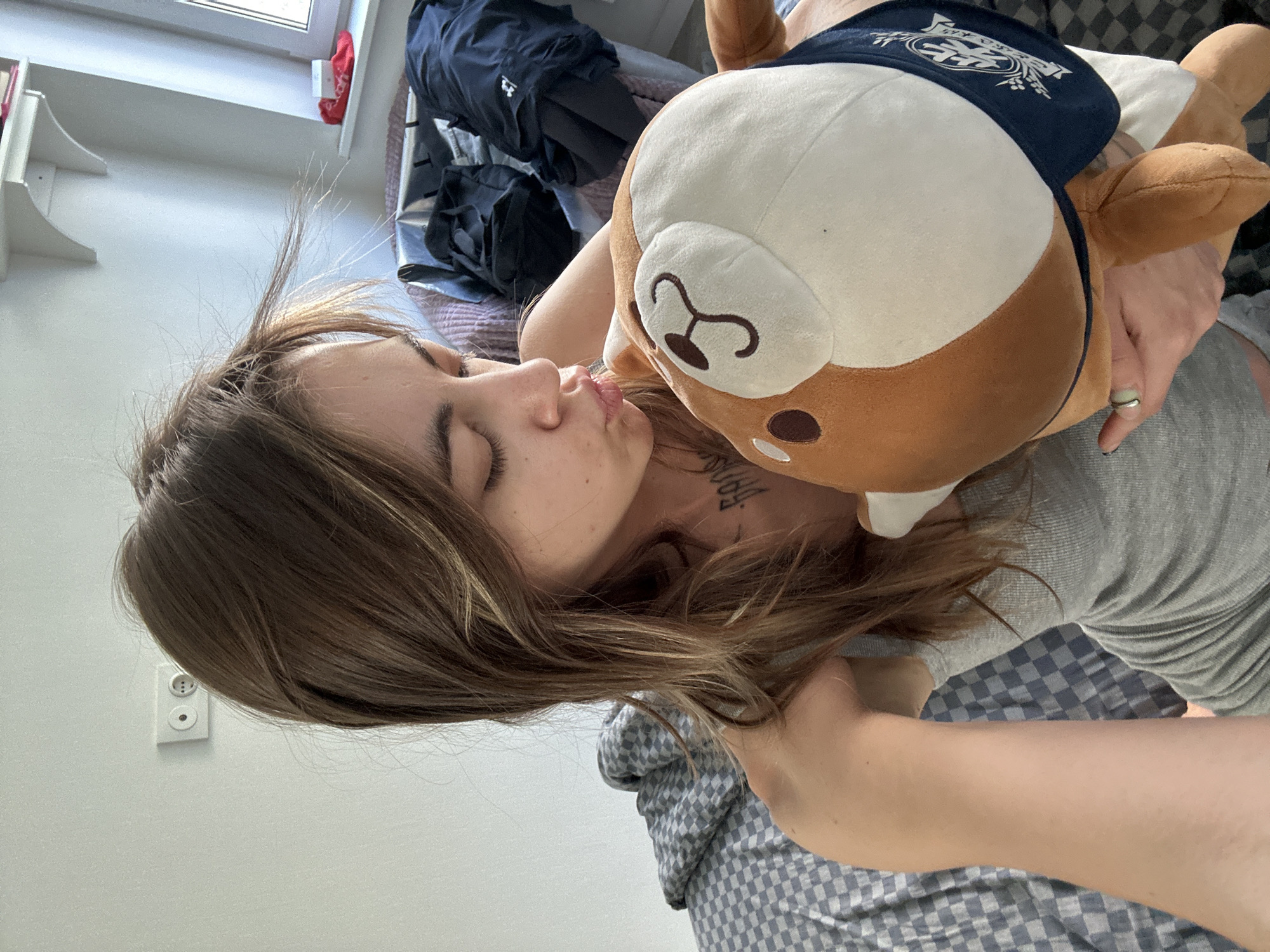 In a brightly lit room, a young woman with light brown hair is captured in a close-up shot, holding a brown and white stuffed animal. She is wearing a grey tank top and is leaning in to kiss the stuffed toy, her eyes closed. The background reveals a checkered grey and white comforter on a bed, a white wall with an electrical outlet, and a shelf with books and a red item. To the right, a window is visible, and laundry bags filled with dark clothing are piled up. The scene suggests a casual and personal moment within a domestic setting.