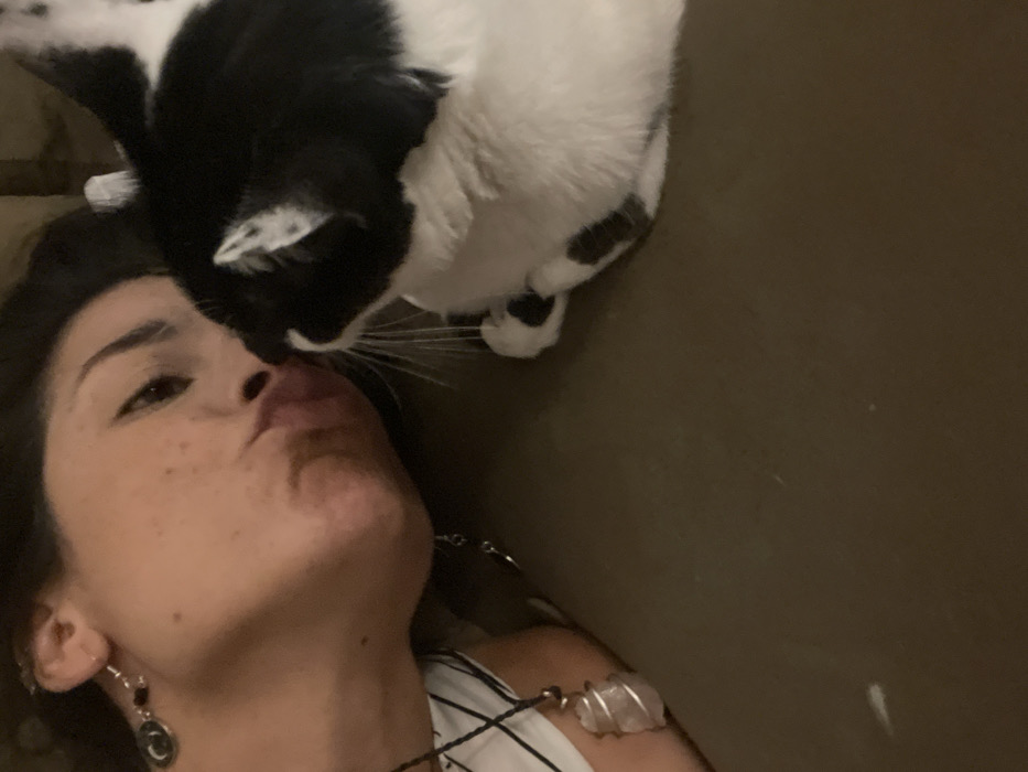 A woman lies on a brown surface with her face angled upward. She wears a white top and jewelry including earrings and a necklace with a clear stone. A black and white cat hovers close to her face, seemingly near her mouth. The cat is positioned above her, with its paws subtly visible. The scene is intimately captured, focusing on the connection between the woman and her pet.