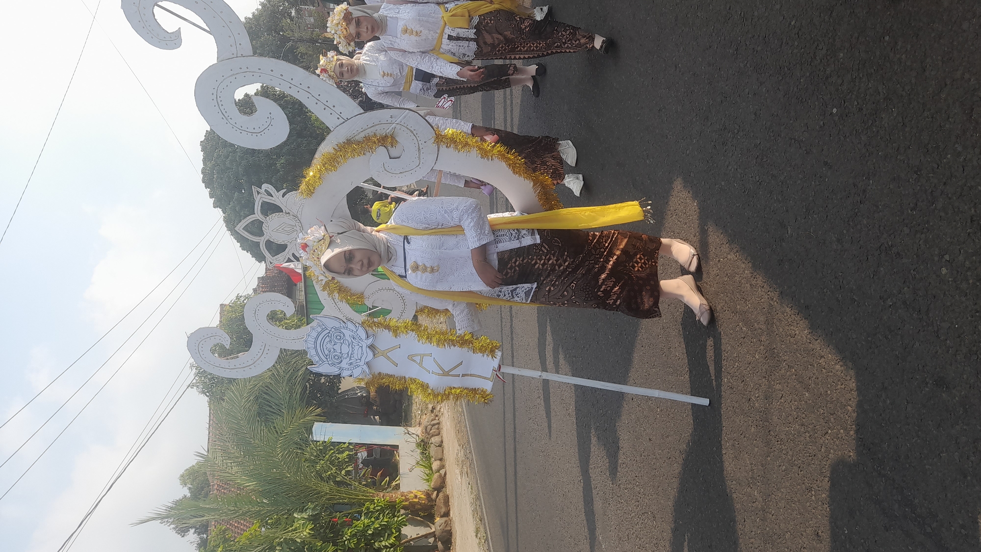 The image captures a daytime outdoor scene on a paved road in Lemahabang, Indonesia. A group of women dressed in traditional Indonesian attire, specifically kebaya and batik, are participating in what appears to be a parade or cultural event. The woman in the foreground is holding a large, white, decorative banner with gold trim, adorned with a mask-like figure and the letters "XAKL." She is wearing a white lace kebaya, a patterned batik skirt, and a bright yellow sash draped over her shoulder. Her head is covered with a light-colored hijab, and she wears a floral headdress. Behind her, two other women are similarly dressed, also wearing floral headdresses. The background shows a residential area with houses and lush green trees under a clear sky with visible power lines. The sun casts long shadows on the road, indicating it is likely morning or late afternoon.
