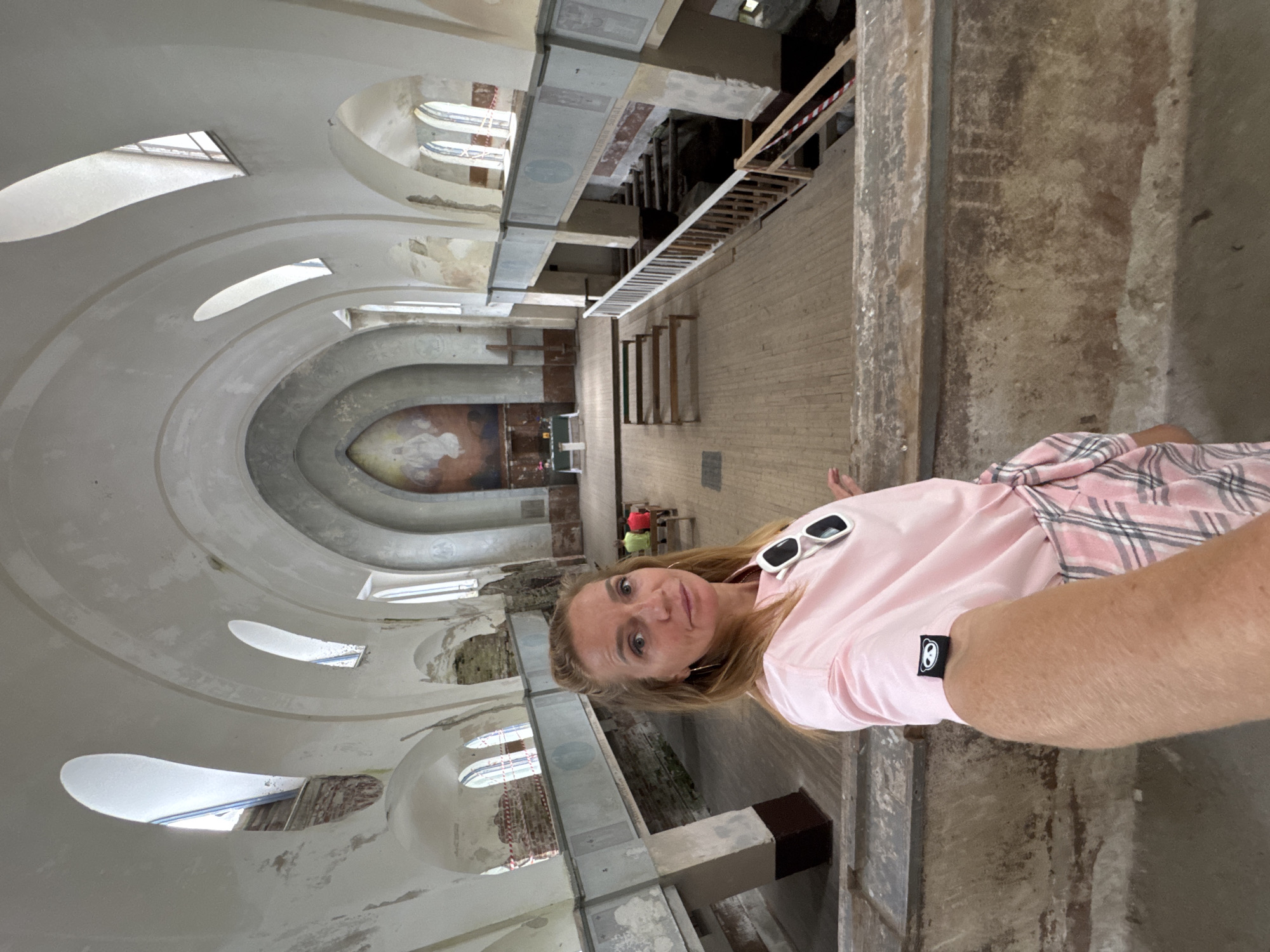 A woman in a pink t-shirt and plaid skirt takes a selfie in an abandoned church in Lumivaara, Russia. The church features arched windows, a fresco on the far wall, and wooden pews. Some of the interior is unfinished, with exposed brick and wooden beams visible in some sections.