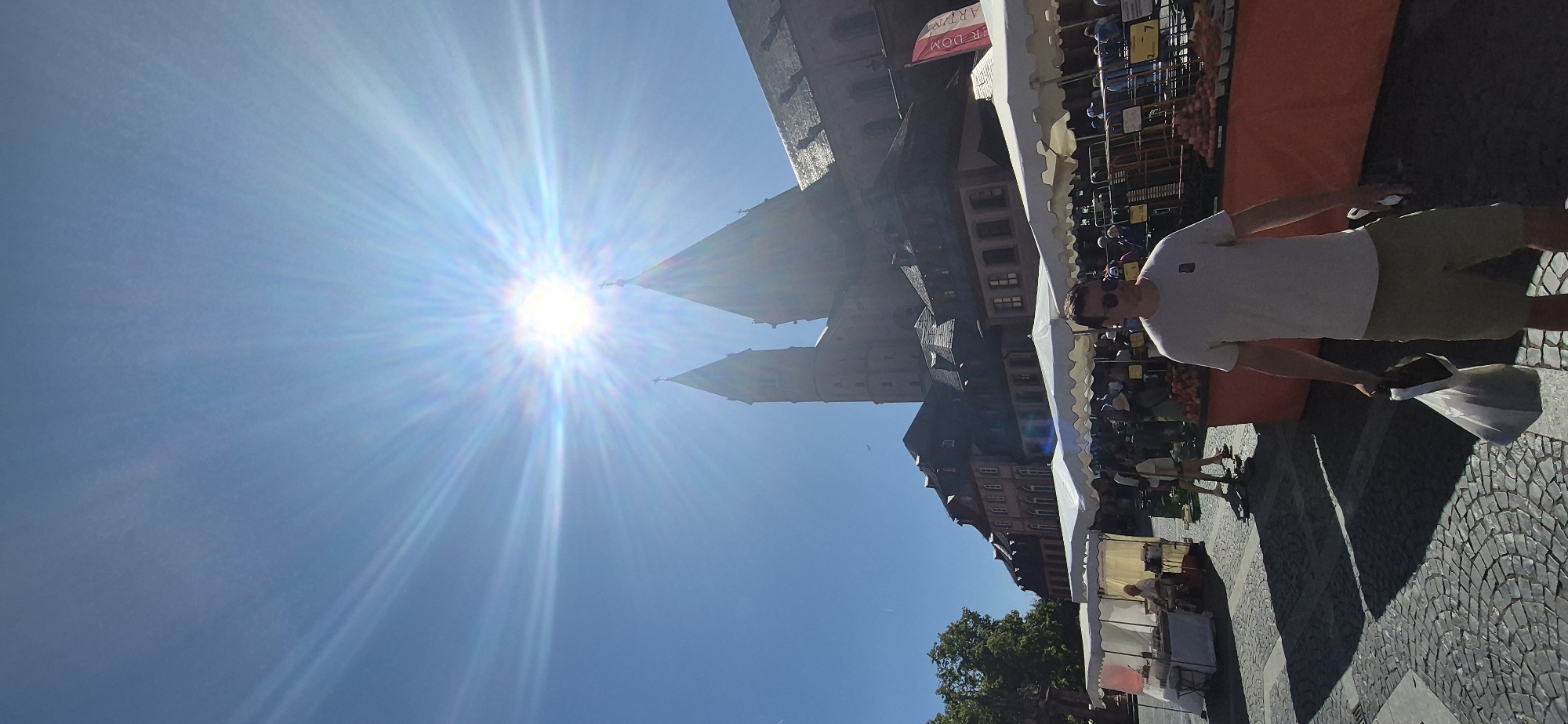 The image depicts a bright, sunny day at a market in Mainz, Germany. In the background, the prominent structure of Mainz Cathedral (Mainzer Dom) is visible, its spires reaching towards a clear blue sky. The sun is high and bright, creating a lens flare effect that radiates across the sky.
In the foreground, a man wearing a light-colored t-shirt, khaki shorts, and sunglasses walks from right to left, carrying a white plastic bag in his left hand. He is walking on a cobblestone street.
The middle ground of the image features market stalls with white canopies. People can be seen browsing the stalls. One stall on the left displays food items. A red barrier or stall front is visible on the right, with produce, possibly tomatoes, arranged on it. A red and white flag with text that appears to be "Dom" and "Martini" is affixed to a pole behind the stalls on the right.
The overall scene captures a lively outdoor market setting in front of a historic landmark on a sunny day.