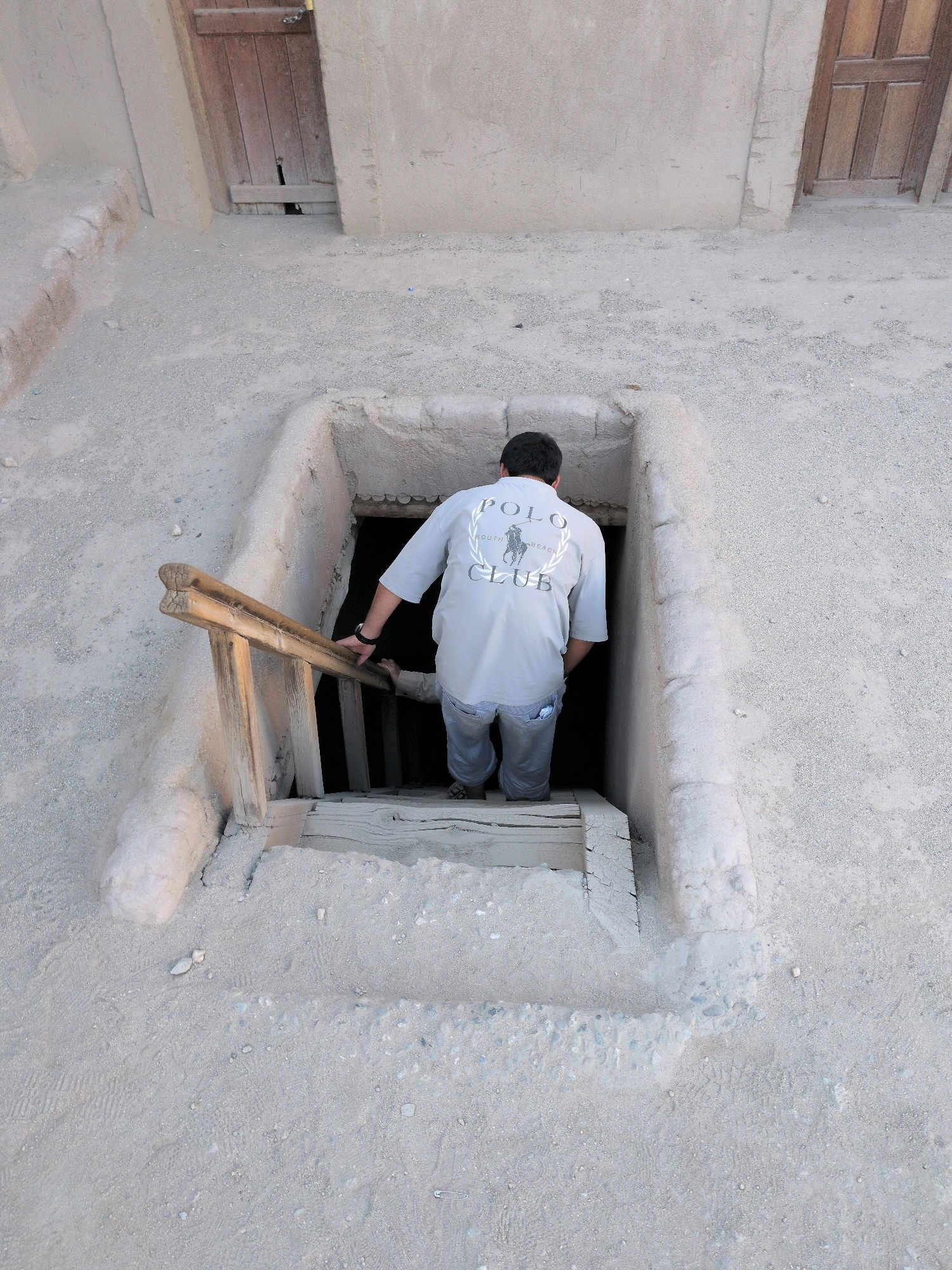 A man wearing a light gray Polo Club t-shirt and blue jeans is descending into a dark opening in the ground. He is holding onto a wooden railing that appears to be part of a makeshift staircase. The opening is surrounded by rough-hewn earth walls. The ground surrounding the opening is sandy and appears to be outdoors in a dry, possibly desert-like environment. The man is seen from behind, with his head and torso angled downwards as he navigates the steps. The Polo shirt has a circular logo on the back. Wooden planks are visible as part of the steps leading into the opening. The overall impression is one of entering an underground structure or passage in a rustic, possibly historical setting. The scene does not provide enough information to determine a specific location in Leh, India.