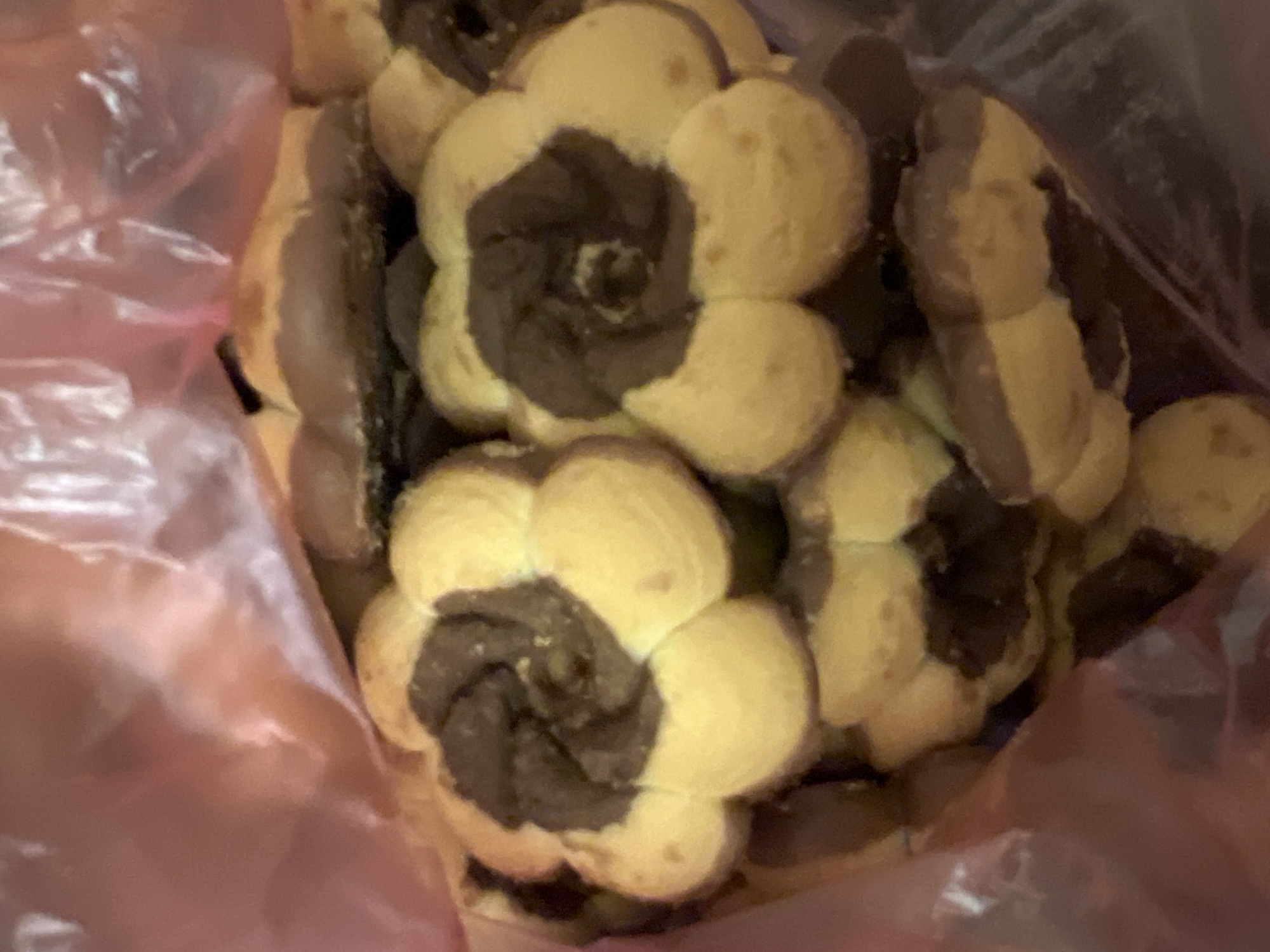 A close-up, overhead shot shows a pile of flower-shaped cookies, some with two-tone colors of light yellow and dark brown, inside a clear plastic bag. The bag is pinkish due to a light source. The cookies have a textured, swirled pattern in their brown centers. The scene is devoid of any geographical indicators placing it in Tolyatti, Russia, or any other specific location. The image primarily focuses on the food item, with no people, actions, or interactions depicted. The background is obscured by the plastic bag and the cookies themselves.