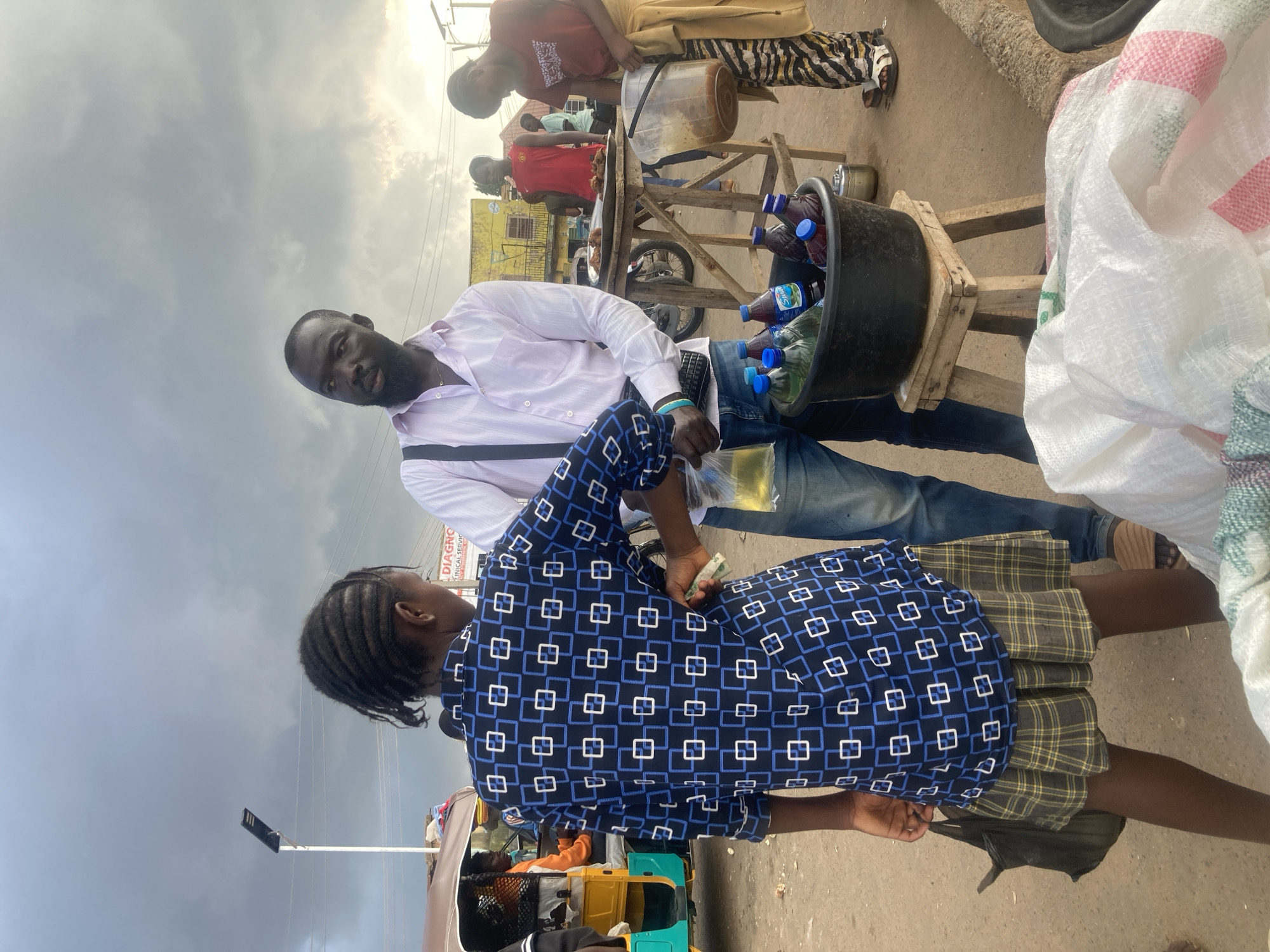 A group of people are gathered on a street in Gwong, Nigeria, with a cloudy sky overhead suggesting it is daytime. In the foreground, a man wearing a light pink shirt and blue jeans is speaking with a young woman dressed in a blue and white patterned top and a plaid skirt. The woman appears to be handing money to the man.
Behind them, a street vendor's stall is set up with various items for sale. A black tub filled with plastic bottles of drinks sits on a wooden stand. Further back, another vendor is tending to food items displayed on a wooden table, with a large clear container of what appears to be palm oil or cooking oil. Several other individuals are visible in the background, some seemingly customers and others assisting the vendors. The scene suggests a busy marketplace or roadside vending area. The overall mood seems neutral, reflecting a typical day of commerce. No explicit text is legible in the image.