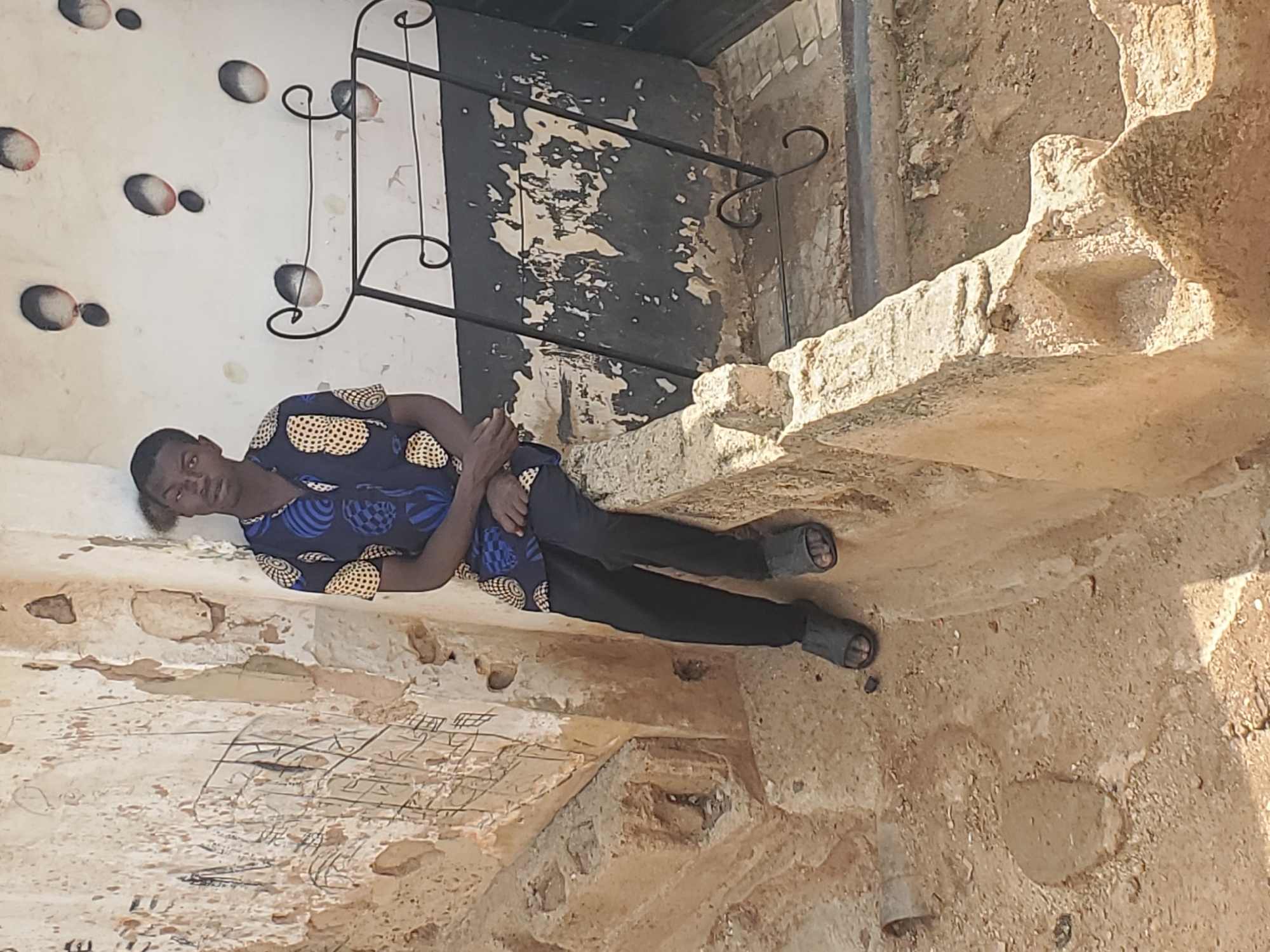 The image shows a young Black male sitting on a low concrete wall outdoors. He is wearing a dark blue short-sleeved shirt with a yellow and white circular pattern, and black pants with black sandals. His left arm is resting on his lap and his right arm is bent, with his hand resting on his knee. The background consists of a weathered, textured wall with some black and white paint peeling off. There is a black metal decorative railing behind him, and the ground is covered in dirt and sand. The wall has some black scribbles on it, possibly graffiti. The overall setting appears to be an urban environment in Gwong, Nigeria, likely a residential or public outdoor space.