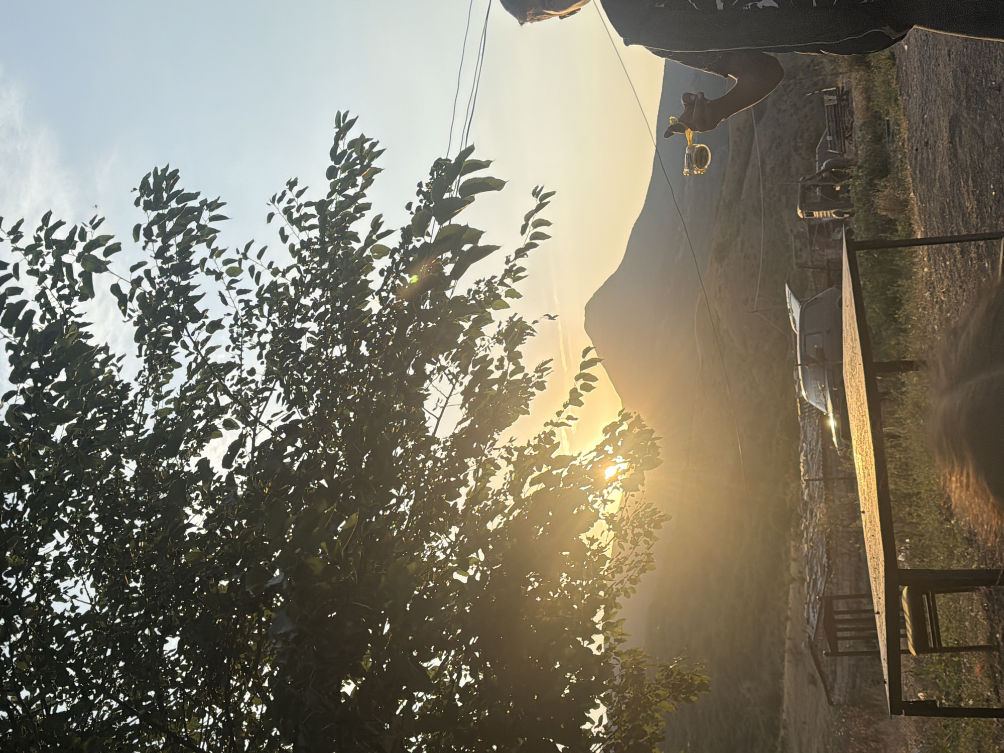 An outdoor scene captures a person's right hand holding a slingshot, with a yellow band and a small projectile, against a backdrop of rolling hills and a hazy sky. The sun is setting, casting a warm glow and lens flare through the leaves of a large tree in the foreground, partially obscuring the view of the landscape. In the middle ground, a wooden table and a chair are visible, along with several vehicles, including what appears to be a white van and a dark-colored truck or SUV. The location is identified as Köhnə Quşçu, Azerbaijan. The overall impression is one of a rural, possibly arid environment during a golden hour sunset.