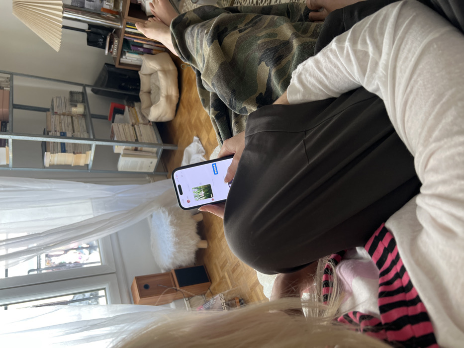 This image depicts an indoor scene within an apartment in Paris, France. The perspective is an over-the-shoulder shot of a person, likely a woman with blonde hair, seated and looking at a smartphone. She wears a white long-sleeved top with a pink and black striped cuff visible on her left arm. Her hands hold the black smartphone, whose screen displays an image of green foliage, likely a plant, with application interface elements visible.
To the right of the foreground person, a second person's lower body is partially visible; their bare feet are resting, and their legs are covered by dark trousers and a camouflage-patterned blanket or garment.
The room features a warm, lived-in atmosphere. A large window with white sheer curtains dominates the left side, revealing a glimpse of typical Parisian architecture outside, including a stone building facade and a dark wrought-iron balcony railing. Below the window, two stacked wooden speakers or audio units sit on the hardwood floor next to a small, white, furry stool with three light-colored wooden legs.
Against the far wall, a tall, multi-shelf metal unit is densely packed with numerous books, both vertically and horizontally stacked. Below this unit, a beige pet bed rests on the wooden floor. To the right of the metal unit, a floor lamp with a pleated shade and a smaller wooden shelving unit, also filled with books and various decorative items, are visible. An older model television screen can be seen on a lower shelf of the metal unit. The room is illuminated by natural light entering through the window.