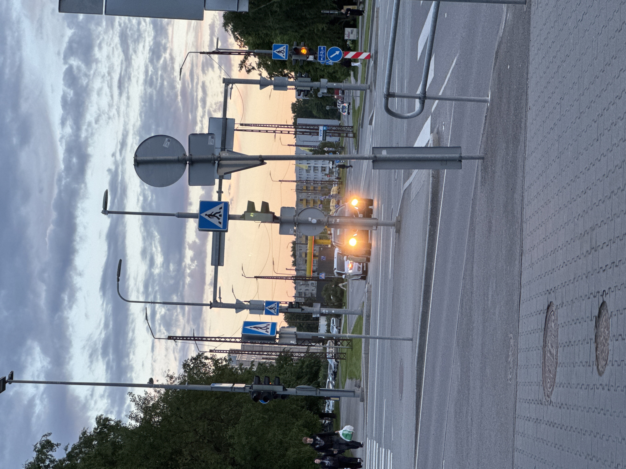 The image depicts a street intersection in Tallinn, Estonia, during twilight. The sky is overcast with clouds tinged by the setting sun. Several traffic lights are visible, with one displaying a yellow signal. Pedestrian crossing signs, depicting a person walking, are present on poles. A few vehicles are on the road, including a car with its headlights on. Two individuals are walking on a paved sidewalk, one carrying a shopping bag. Street lamps are illuminated, providing light. Buildings and trees form the background of the scene. The intersection includes road markings for pedestrian crossings and lanes.