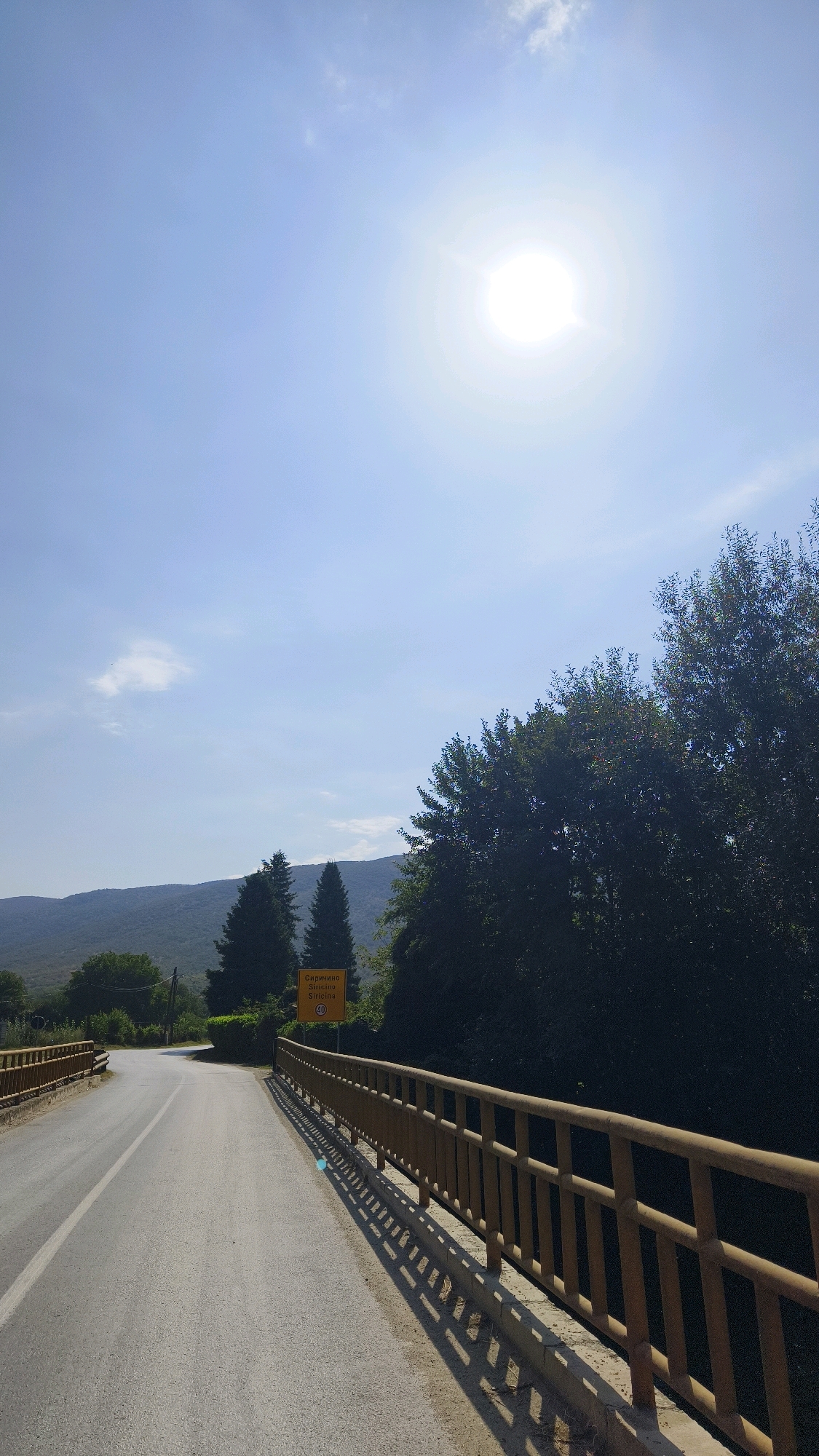 The image depicts a road leading over a bridge in Sirichino, North Macedonia, under a bright, sunny sky. The road curves to the left and is bordered by a wooden guardrail on the right side of the frame. Dense green foliage and trees are visible on the right and in the background, partially obscuring the mountainous landscape. A yellow road sign with text and a speed limit of 40 is positioned on the left side of the road near the bridge entrance. The sun is prominently featured in the upper right quadrant of the sky, casting shadows from the guardrail onto the road. The scene appears to be a rural or semi-rural setting during the daytime.