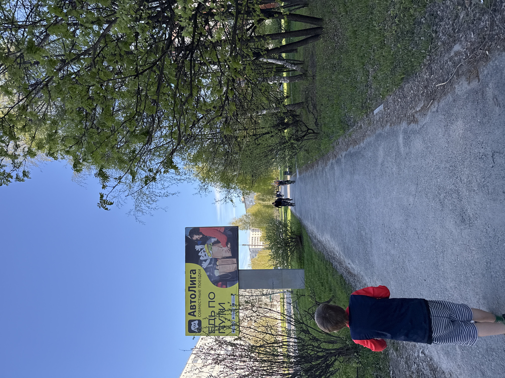 The photograph captures a scene from what appears to be a pedestrian walkway in Tobolsk, Russia, characterized by a tree-lined path and a bright, clear sky.
A young boy is visible in the foreground with dark striped shorts, a blue t-shirt, and red sleeves. He is positioned at the side of the walkway, his back facing the camera.
Along the pathway, there are two individuals on bicycles moving away from the viewer. Trees flank both sides of the walkway, with the walkway itself being composed of gray gravel.
A billboard is situated along the path with Russian writing and an advertisement. Behind the billboard are some buildings.