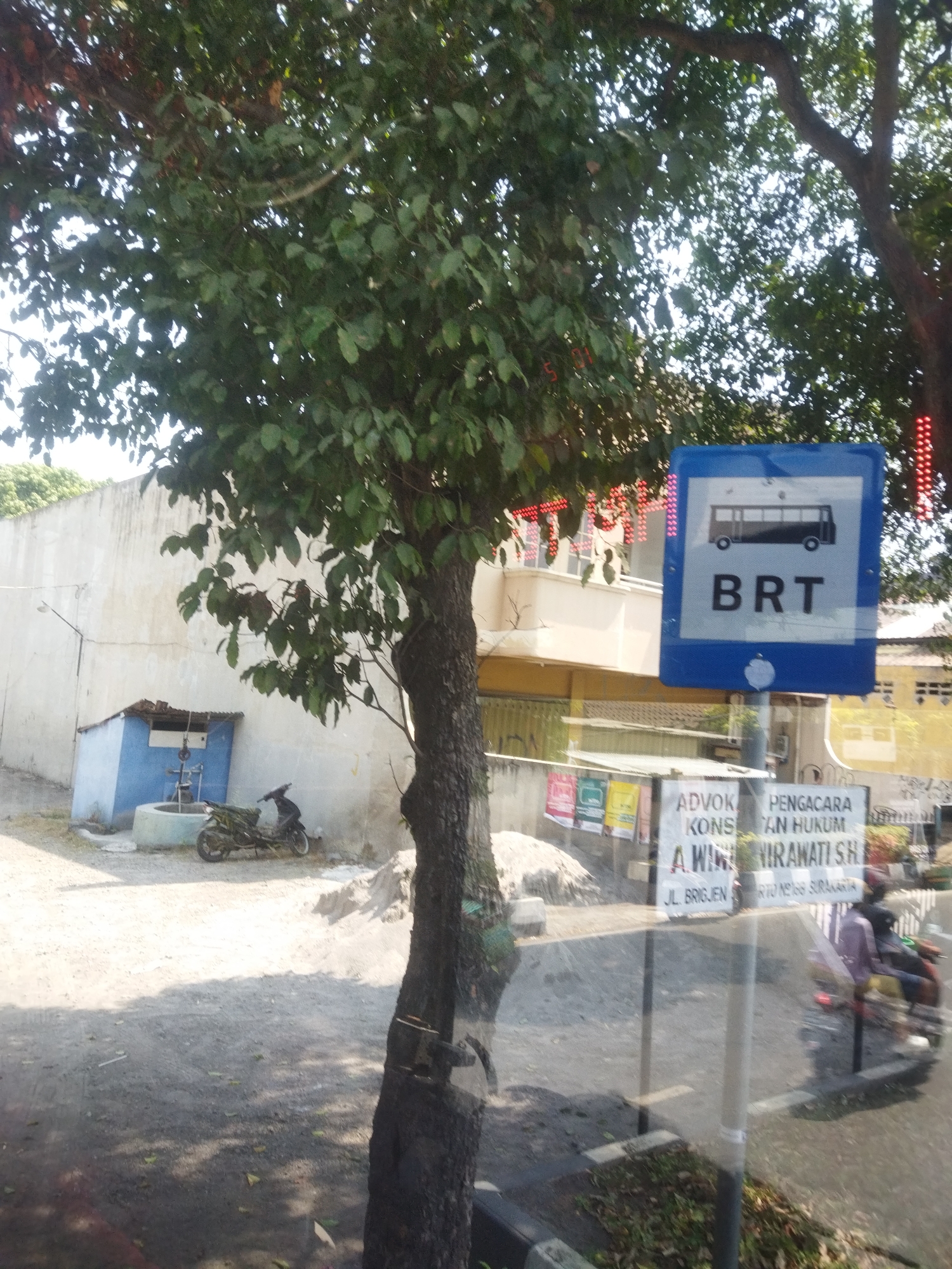 The image captures a street scene in Surakarta, Indonesia, with a prominent BRT (Bus Rapid Transit) bus stop sign on the right. A large tree with dense green foliage partially obscures the view on the left. In the background, a light-colored building with a tiled roof stands next to a smaller, blue structure with a well. A parked motorcycle is visible in front of the blue structure. Further to the right, a blurred image shows people on a motorcycle. Several signs are posted, including one that reads "ADVOC PENGACARA KONSULTAN HUKUM A. WIRAWATI SH" and another with an address that appears to be "JL. BRIGJEN" and "RTQ NO.166 SURAKARTA". The ground is unpaved and dusty. The lighting suggests it is daytime, with sunlight casting shadows.