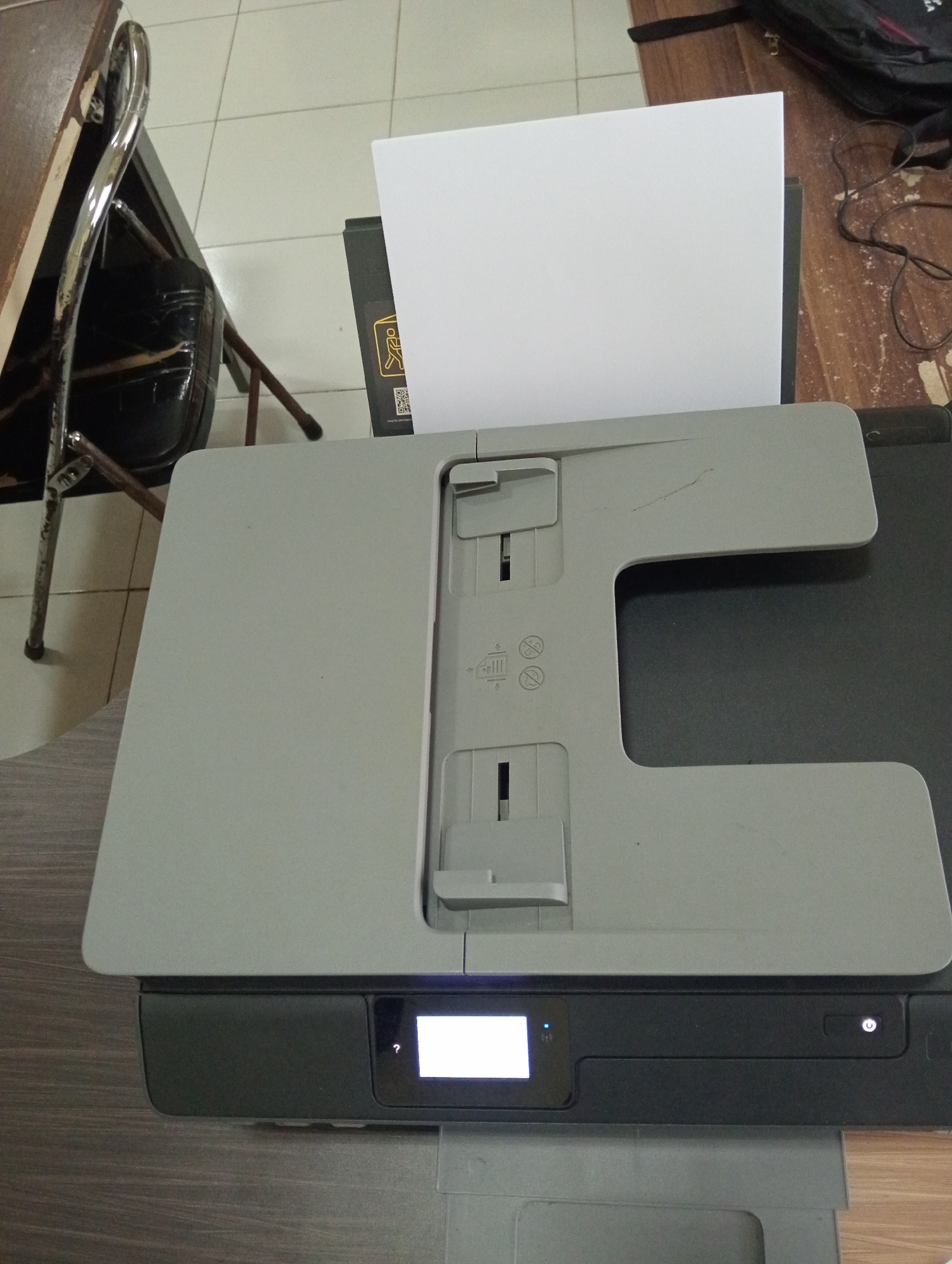 An overhead, close-up, static shot captures a portion of a printer on a wooden desk. The printer is primarily dark gray and light gray, with a small white touchscreen displaying a white screen and a question mark symbol. The top of the printer features a large, light gray flatbed scanner lid, which is slightly ajar, allowing a white sheet of paper to be visible. The paper is positioned in the document feeder. To the left of the printer, a portion of a wooden table and the chrome legs of a chair are visible. The flooring is tiled in a light color. In the background, a black backpack and some electrical cords are scattered on the wooden floor. The scene is indoors, suggesting a workspace or home office environment. The location is Sidoharjo, Indonesia.