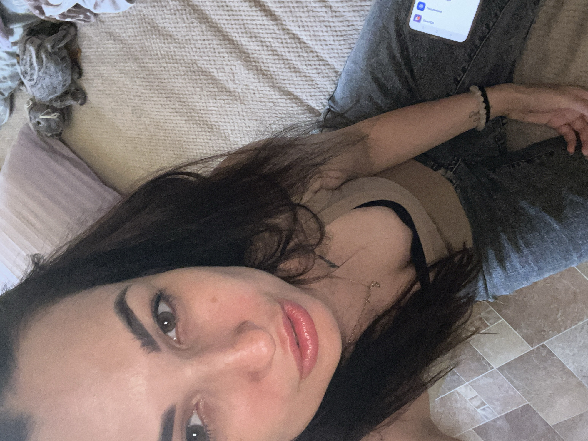 A young woman with dark hair and brown eyes is lying on a bed. She is wearing a beige tank top and blue jeans. She is looking at the camera and smiling. There is a stuffed animal on the bed next to her. The room is dimly lit.