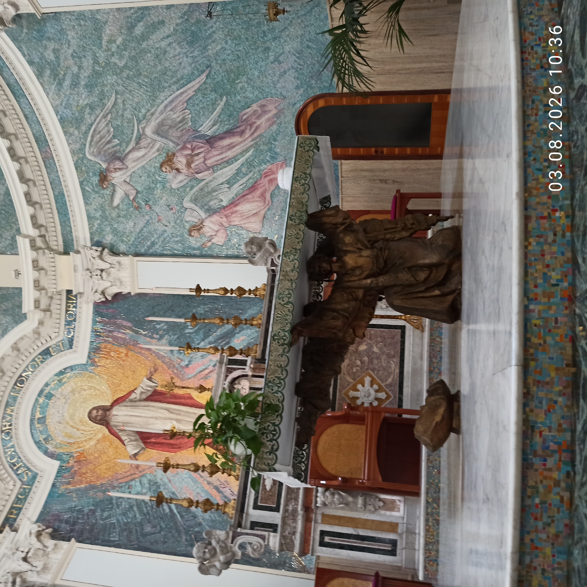 The image depicts the interior of a church, likely in Ruvo di Puglia, Italy, showing an altar area. In the background, a large mosaic adorns the wall, featuring a depiction of Jesus Christ in the center, with angels surrounding him. In front of the mosaic, an altar stands, adorned with white cloth and greenery. Beneath the altar, a bronze sculpture of an angel with outstretched wings is positioned. The floor is made of marble, with a colorful mosaic base on the altar steps. A stone object rests on the floor in front of the altar. The lighting suggests an interior setting, possibly with natural light from unseen sources.
