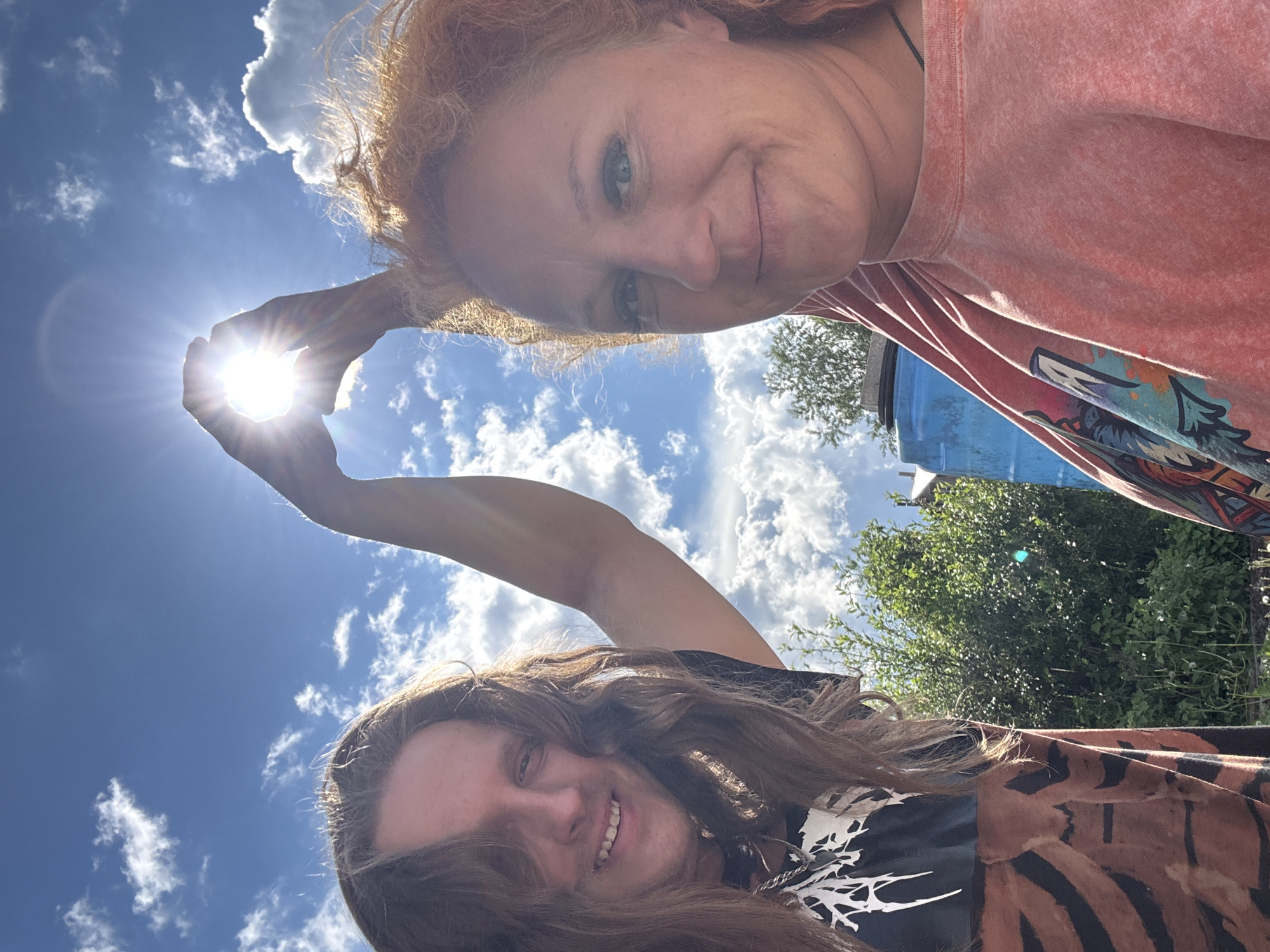 This low-angle selfie features two smiling individuals outdoors under a bright blue sky with scattered white clouds, likely in Chaikovsky, Russia. The camera is positioned looking upwards, creating an inverted perspective where the subjects appear to be looking down at the viewer.
In the upper half of the frame, a person with red or auburn curly hair and light-colored eyes is visible, looking directly at the camera with a gentle smile. They are wearing a peachy-orange t-shirt with a colorful, abstract graphic design.
Below them, another individual with long, wavy dark hair and a wide, bright smile is also looking down towards the camera. They are wearing a black t-shirt with a white graphic pattern and a brown garment with black stripes, possibly an open shirt or blanket.
A notable detail is the playful interaction with the sun. The red-haired person extends an arm upwards, and their hands are positioned to frame the intensely bright sun, making it appear as if they are holding or cupping it. The sun emits strong rays, creating a visible lens flare.
The background features lush green foliage from trees or bushes on the right side, and a portion of a blue barrel or container. The vibrant blue sky and bright sun indicate it is daytime, likely a sunny and clear day. Both subjects exhibit joyful and relaxed expressions, suggesting a pleasant and lighthearted moment. No legible text is visible on their clothing or in the background elements.