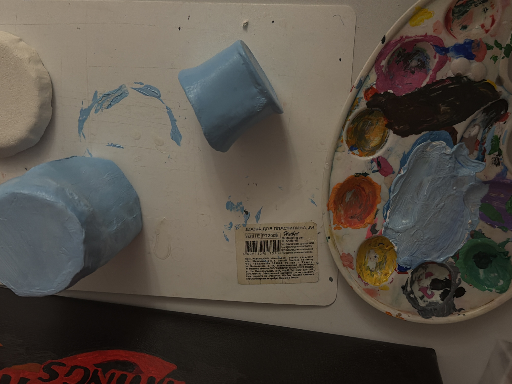 The image shows an art workspace. There's a white plastic board with paint smudges and three light blue, clay-like objects shaped into cylinders. Next to the board is a white painter's palette with various colors of paint including blues, oranges, pinks, greens, and brown. A partial view of what appears to be a painted canvas or board with red lettering is visible at the bottom. The white plastic board features a label written in Russian; the product is "ДОСКА ДЛЯ ПЛАСТИЛИНА, A4 WHITE PT2000 Hatler" (Clay Board A4, White PT2000 Hatler). The label indicates that the product was manufactured by the company “OKA-Plast” in Shilovo district, Ryazan region, and produced for the customer "Berlingo" located in Ryazan, Russia.