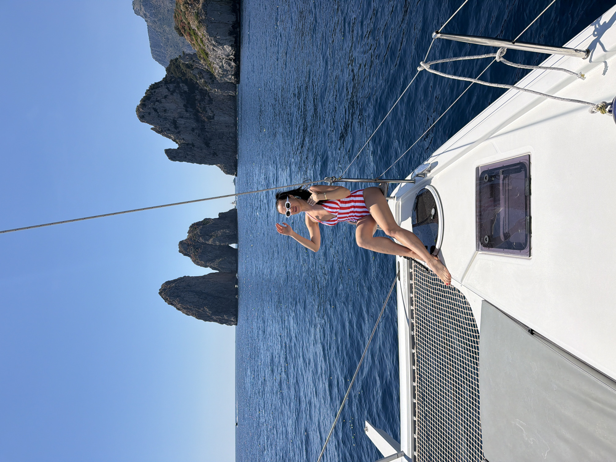 A medium, eye-level shot depicts a woman on a sailboat in the waters off Capri, Italy. She is wearing a red and white striped bathing suit and white sunglasses. The backdrop reveals a deep blue sea and sky. Large rock formations are visible in the background. The woman is positioned on the foredeck of a white sailboat, a netting visible to the side.