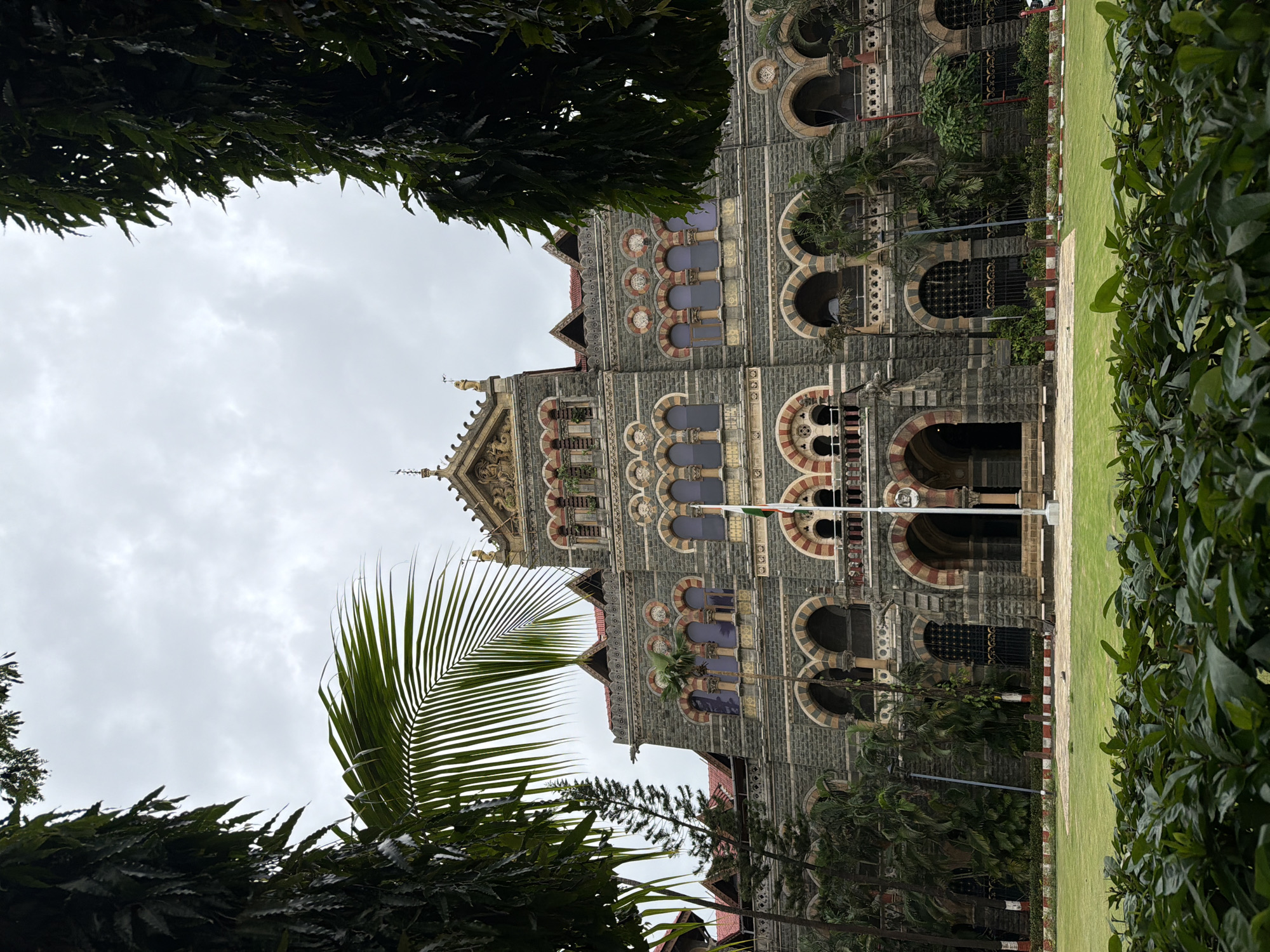 The image shows the Chhatrapati Shivaji Maharaj Terminus, formerly Victoria Terminus, a historic railway station in the Fort, Mumbai, India. The building's Gothic Revival architecture, characterized by its ornate stonework, arched windows, and elaborate towers, is visible. The facade is constructed of red sandstone and blue granite. In the foreground, a neatly mowed green lawn is bordered by a hedge of dark green foliage. A flagpole flying the Indian flag stands in front of the building. The sky above is overcast with thick clouds. Trees with dark green leaves frame the top and sides of the image. There are no people visible in the scene.