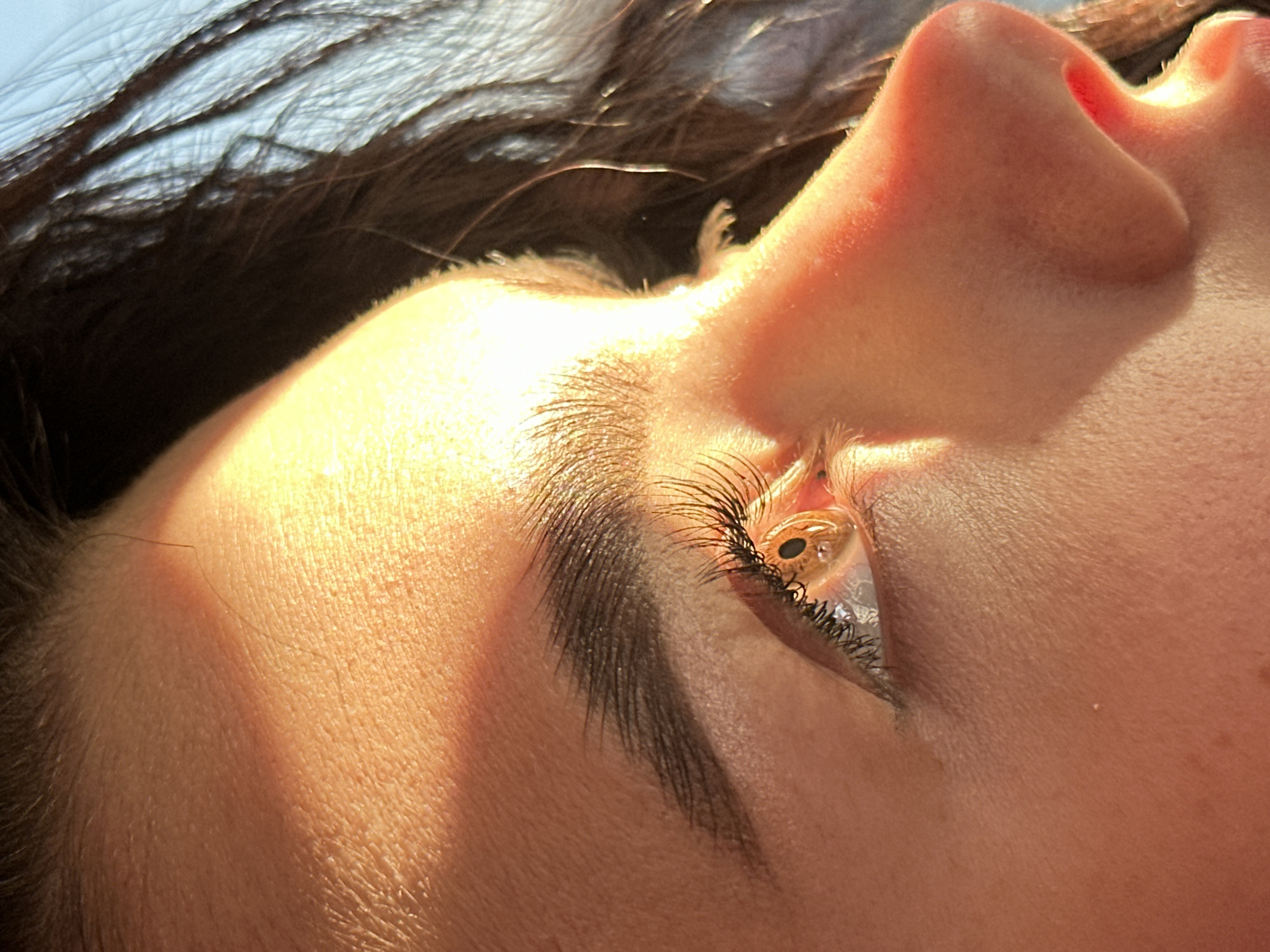 The image is a close-up, upward-angled shot of a person's face, likely a woman, with a portion of dark hair visible in the upper left corner. The subject's face is brightly illuminated by sunlight, casting distinct shadows. The primary focus is on the eye, eyebrow, and nose. The eye is open, with visible eyelashes and a brown iris. The eyebrow is dark and well-defined. The nose is prominently featured, with visible nostril openings. The skin texture is visible, indicating pores and subtle details. The background appears to be a light blue sky, partially obscured by the hair. The location is not discernible from the visual information provided.